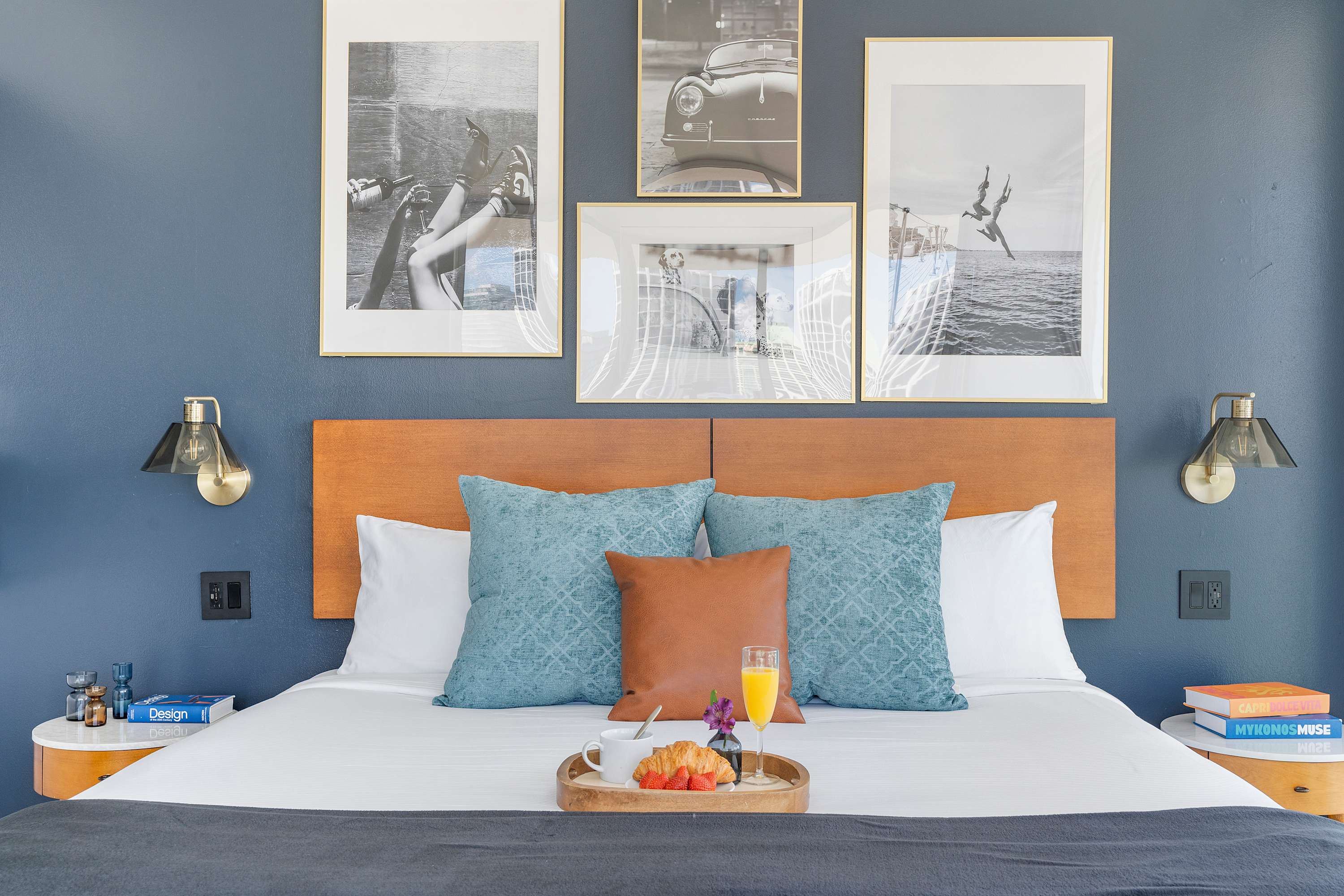 A close-up of the comfortable king-sized bed, adorned with crisp white linens, textured blue throw pillows, and a warm brown leather cushion. The wooden headboard is set against a deep blue accent wall decorated with framed black and white art prints.