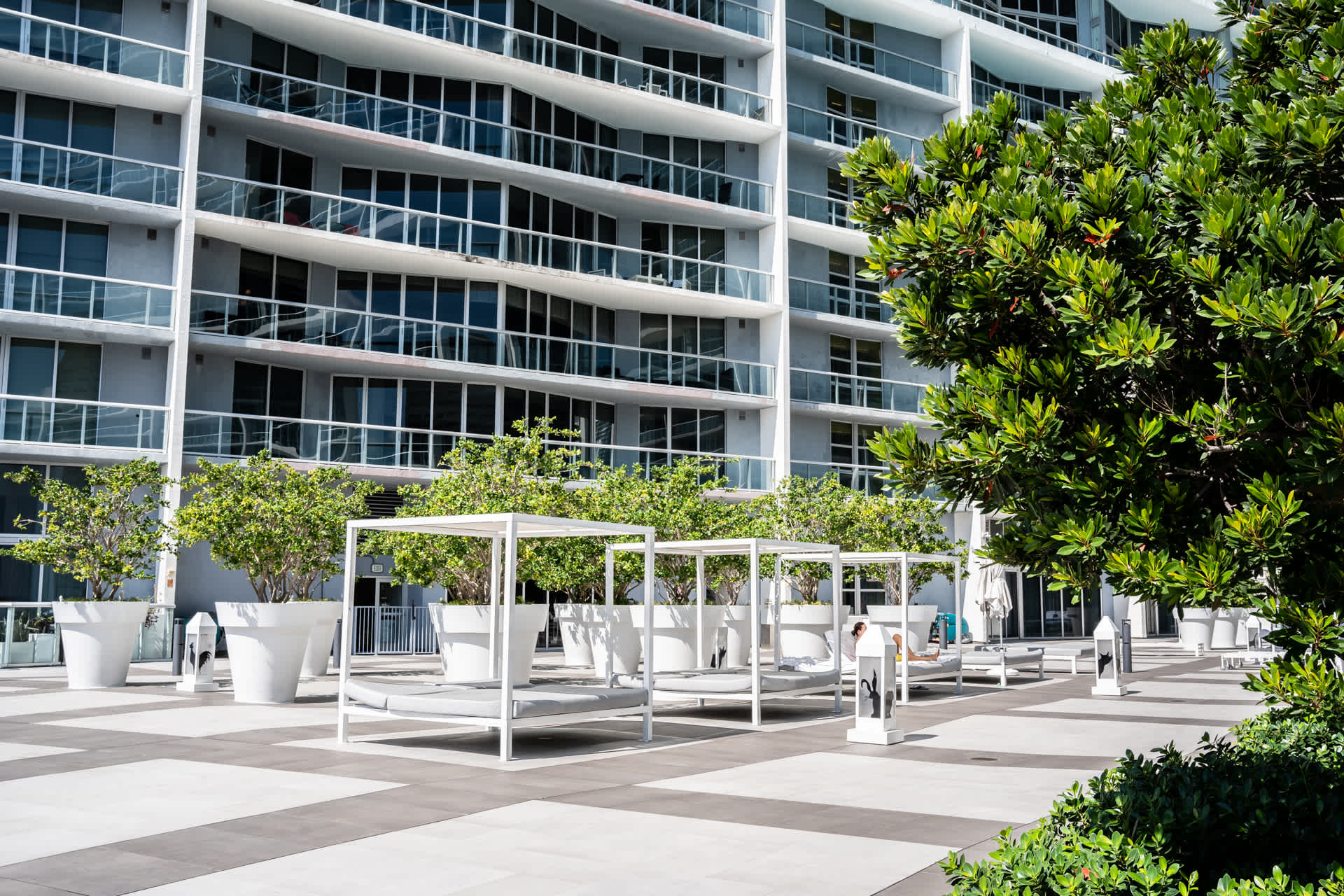 An expansive outdoor pool deck featuring multiple white cabanas and lounge chairs, surrounded by lush green trees and a modern high-rise building in the background.