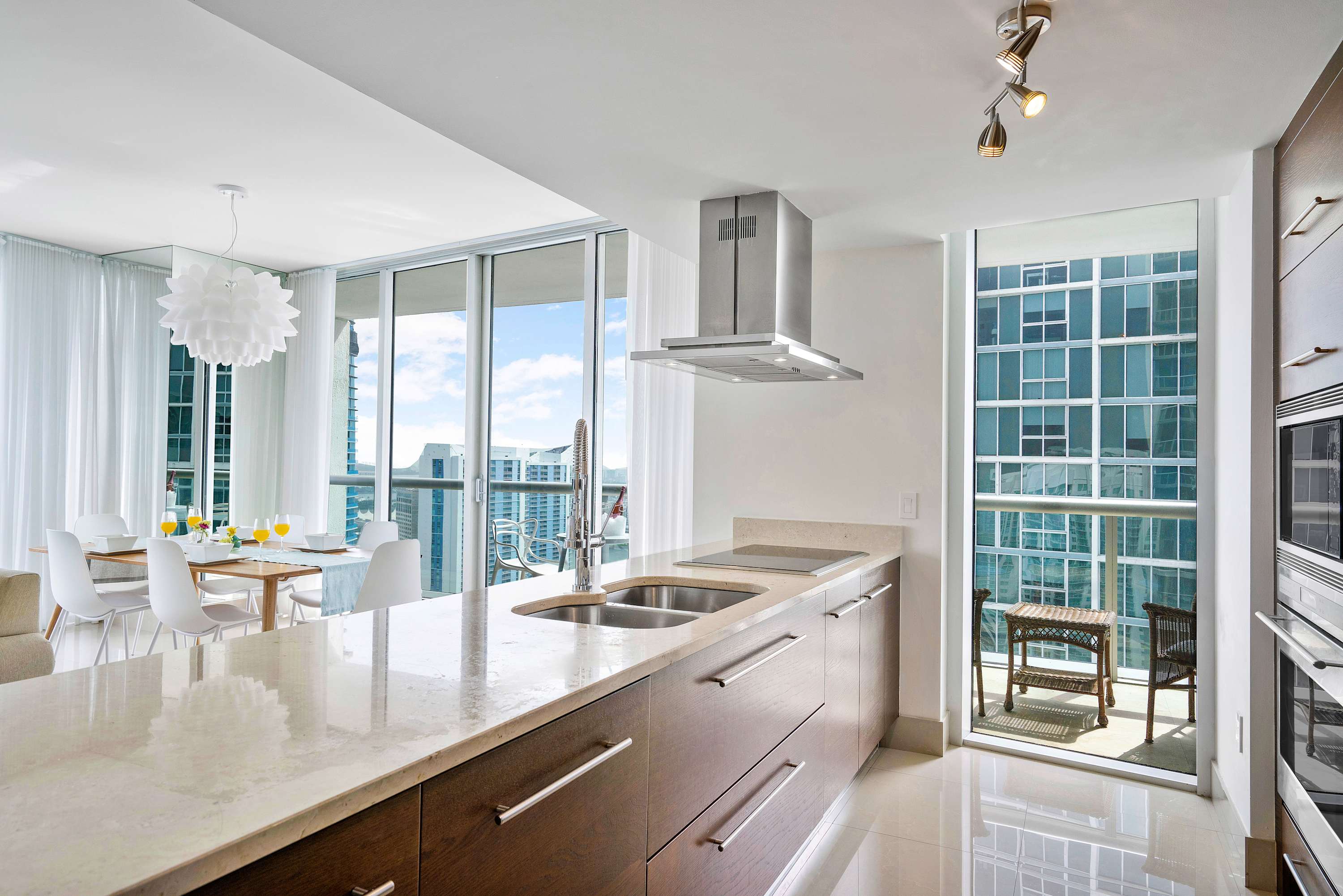 Modern kitchen with light marble countertops, dark wood cabinetry, stainless steel appliances, and a breakfast bar, offering a glimpse of the dining area and a small balcony with city views.