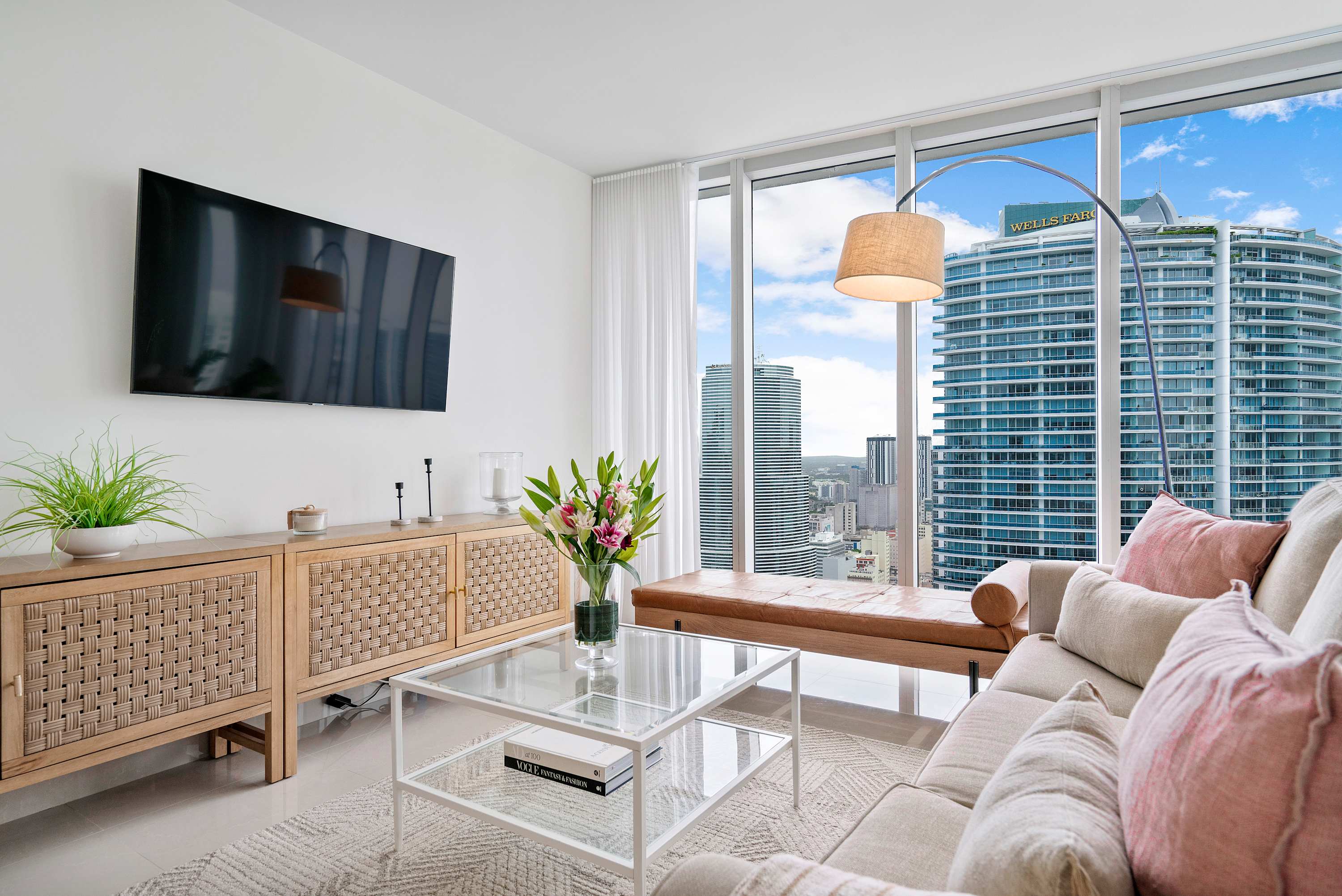 Stylish living room with a large wall-mounted TV above a woven wooden console, a light-colored sofa with pink pillows, and a leather bench, all overlooking a vibrant city skyline through expansive windows.