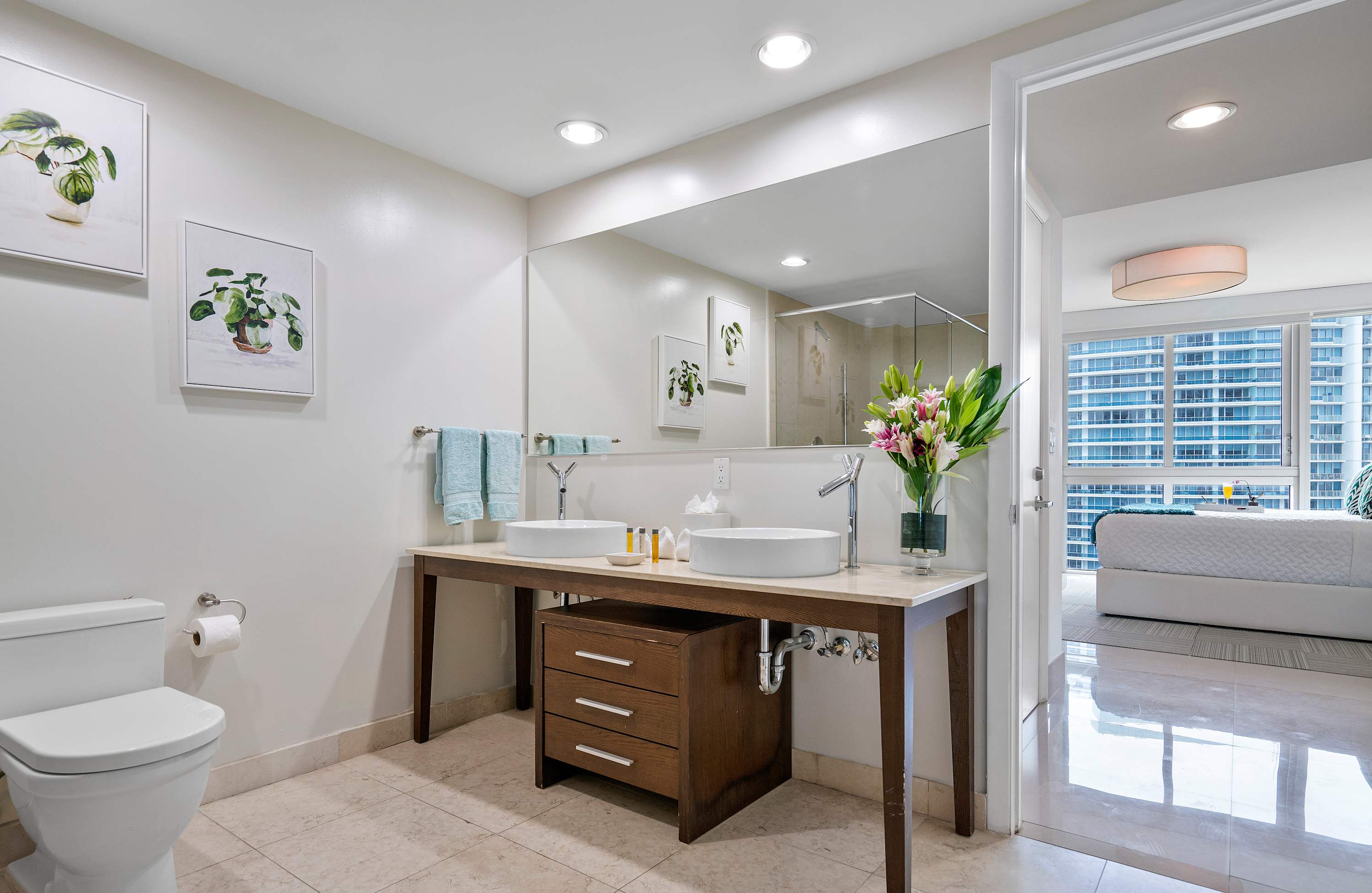 A bright bathroom featuring a double vanity with two white vessel sinks on a light marble countertop, a dark wood base cabinet, and a toilet on the left. A large mirror reflects a glass-enclosed shower and a bedroom with city views.
