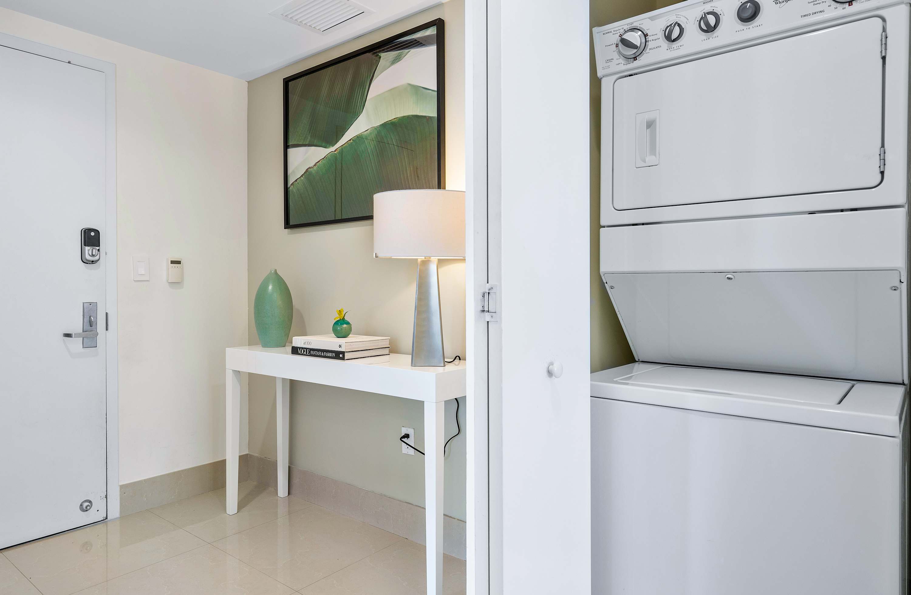 A bright foyer area with light tiled flooring, featuring a white console table adorned with decorative items and a lamp, next to a stacked white washer and dryer unit in a closet. A white door with a smart lock is visible on the left.