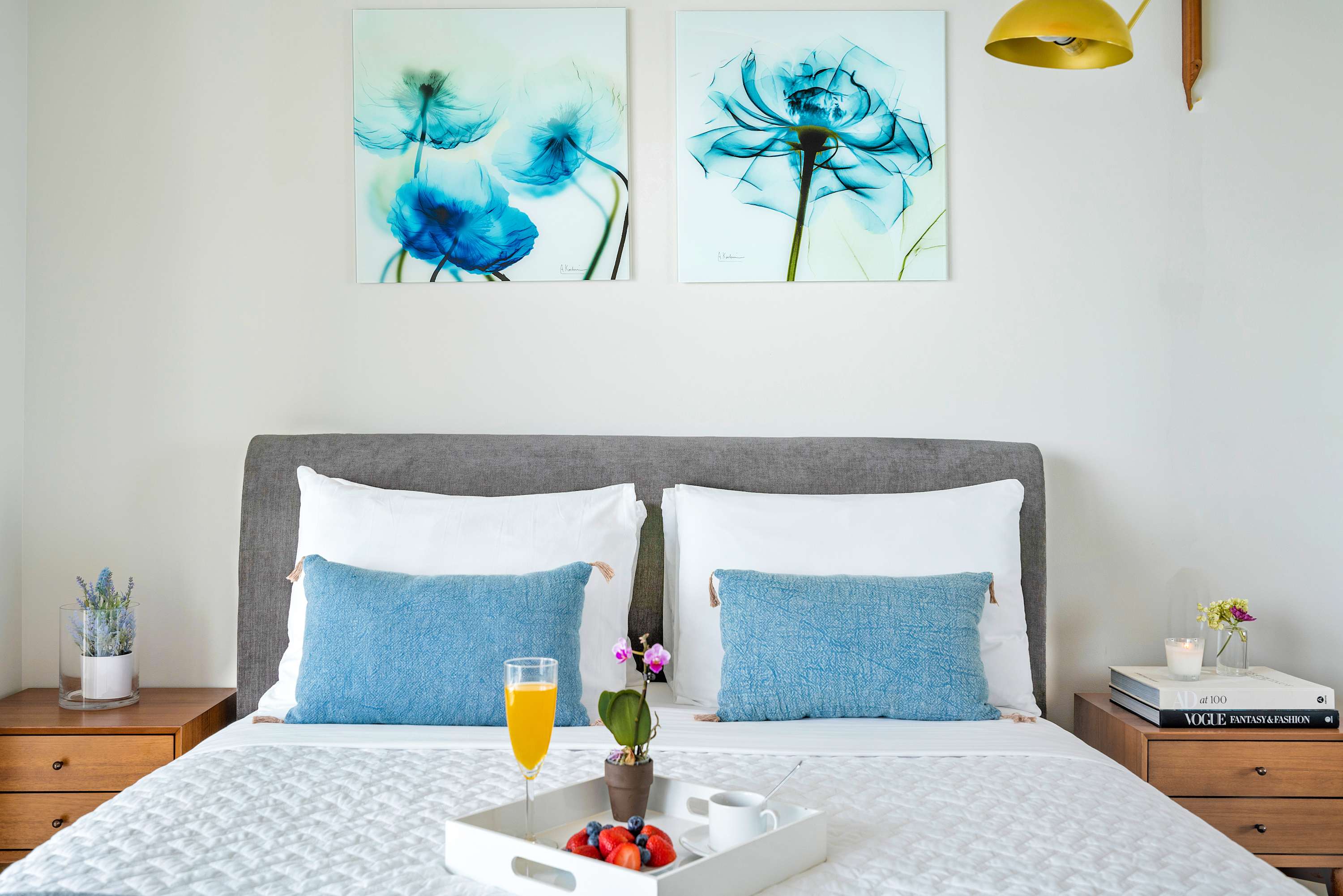Close-up of a stylish bed with a grey upholstered headboard, white bedding, and blue accent pillows, flanked by wooden nightstands with lamps and modern blue floral artwork above.