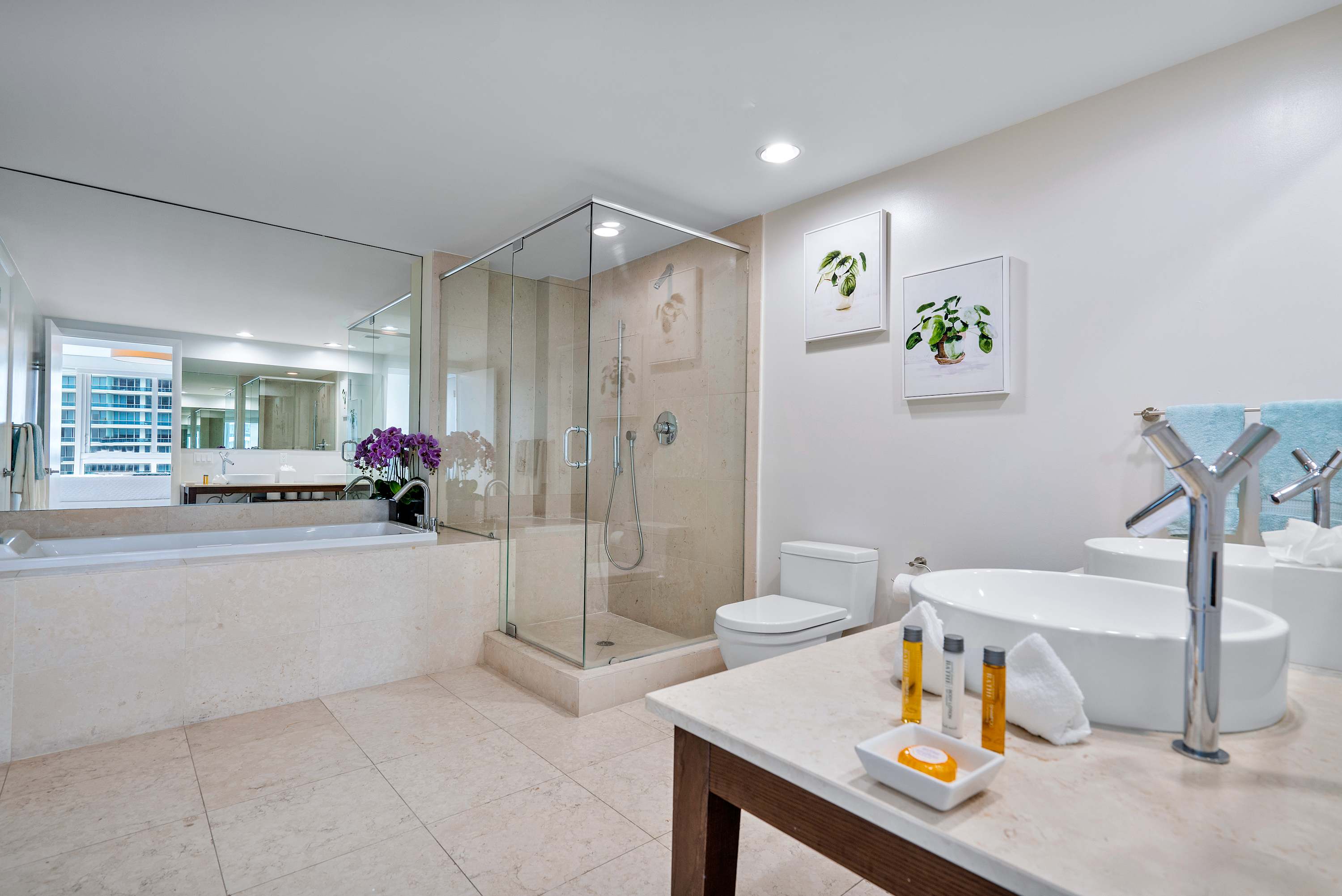 A spacious bathroom with light-colored tiled floors and walls, featuring a glass-enclosed walk-in shower, a built-in bathtub, and a vanity with a single white vessel sink. A toilet is visible next to the vanity.