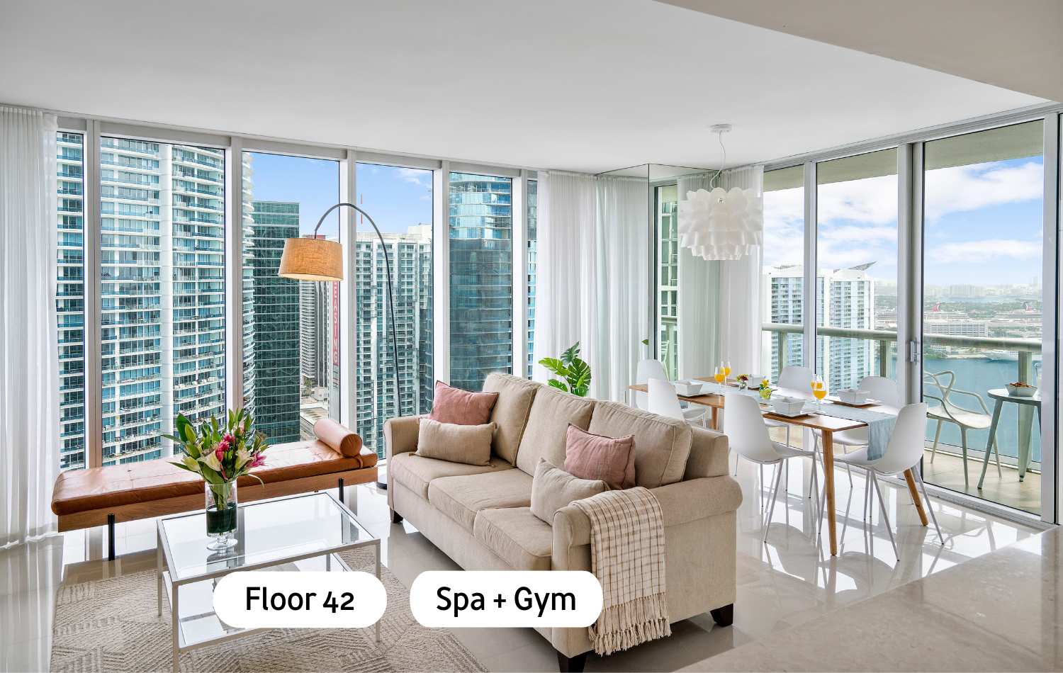 Modern open-concept living and dining area with a light-colored sofa, glass coffee table, and a dining table, all set against floor-to-ceiling windows offering expansive city and waterfront views.