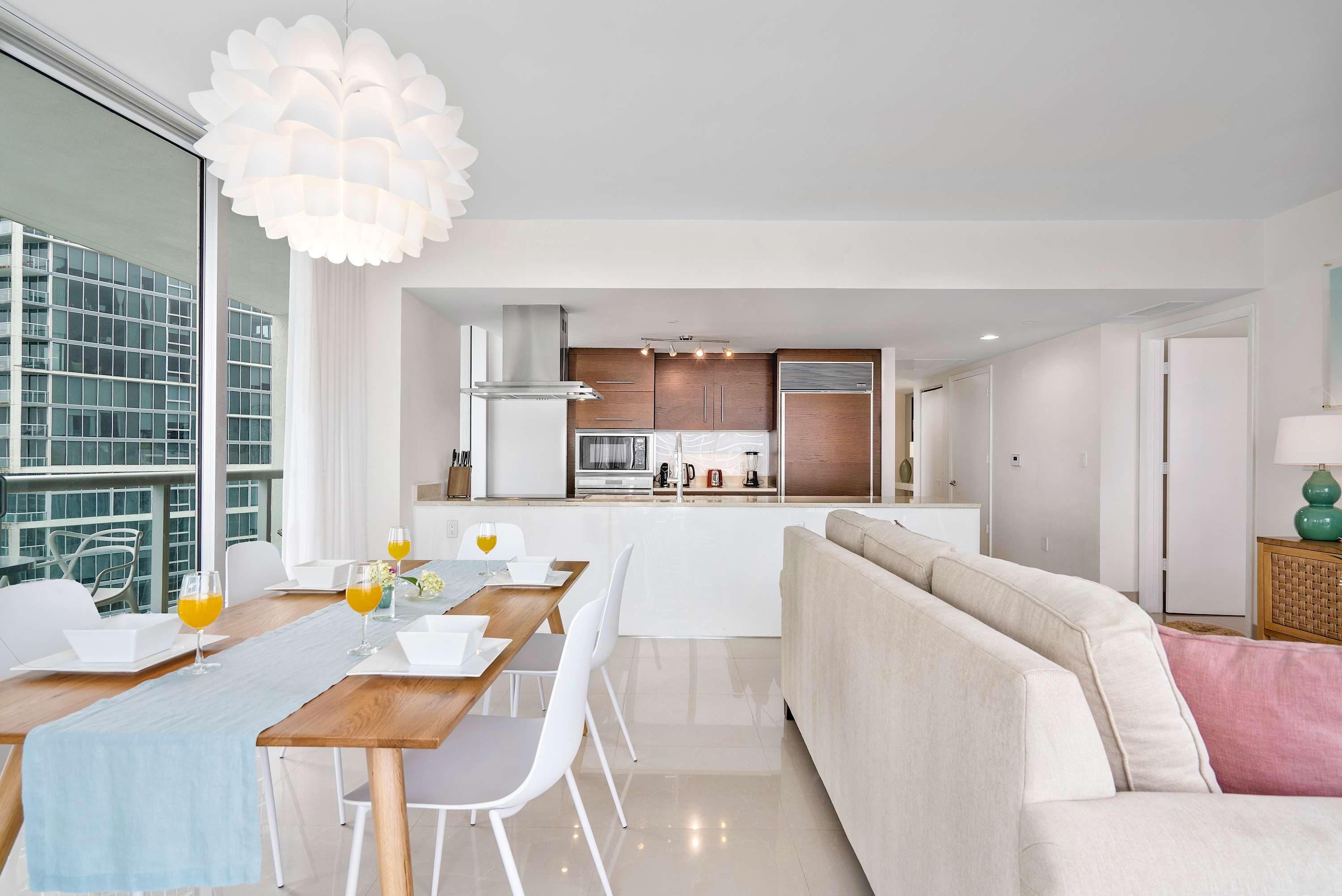 Contemporary dining area with a light wood table and white chairs, illuminated by a large white pendant light, with a modern kitchen visible in the background and city views through large windows.