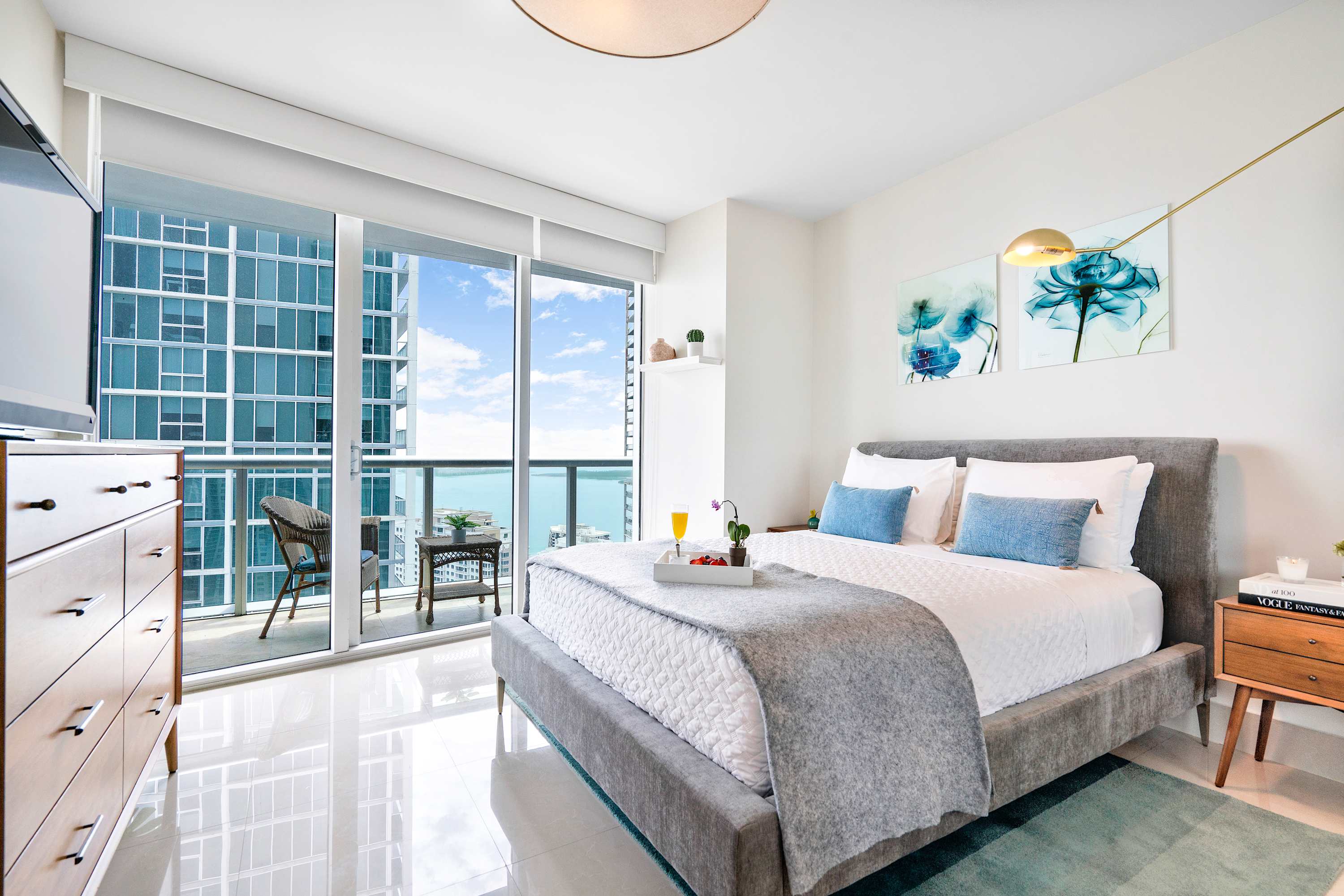 Bright bedroom featuring a grey upholstered bed with blue pillows, a wooden dresser, and a private balcony with seating, offering stunning city and water views.