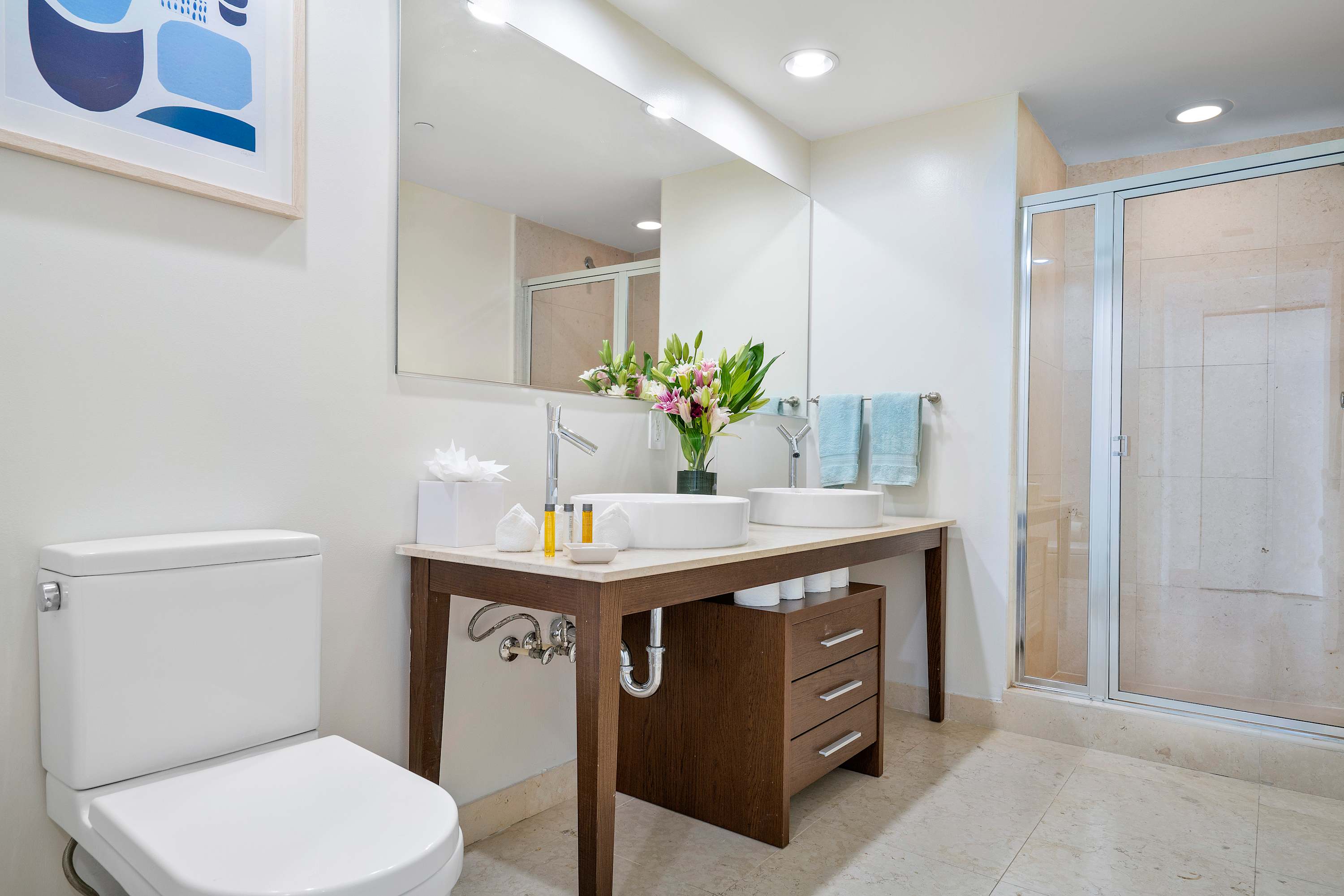 A bright bathroom featuring a double vanity with two white vessel sinks on a light countertop, a dark wood base cabinet, and a toilet on the left. A glass-enclosed shower is visible in the background.