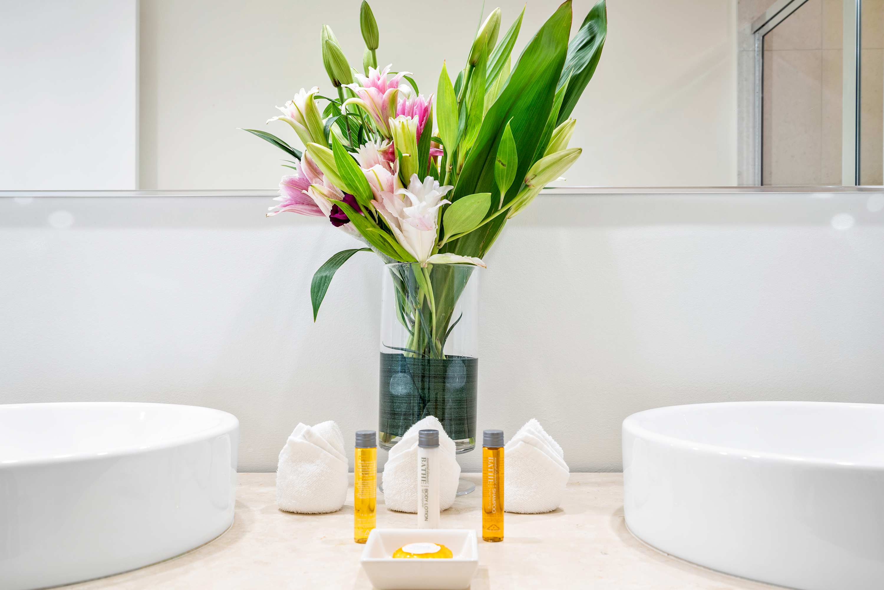 Modern bathroom vanity featuring two white vessel sinks set on a light-colored countertop, with a large mirror above and fresh flowers, along with neatly arranged toiletries.