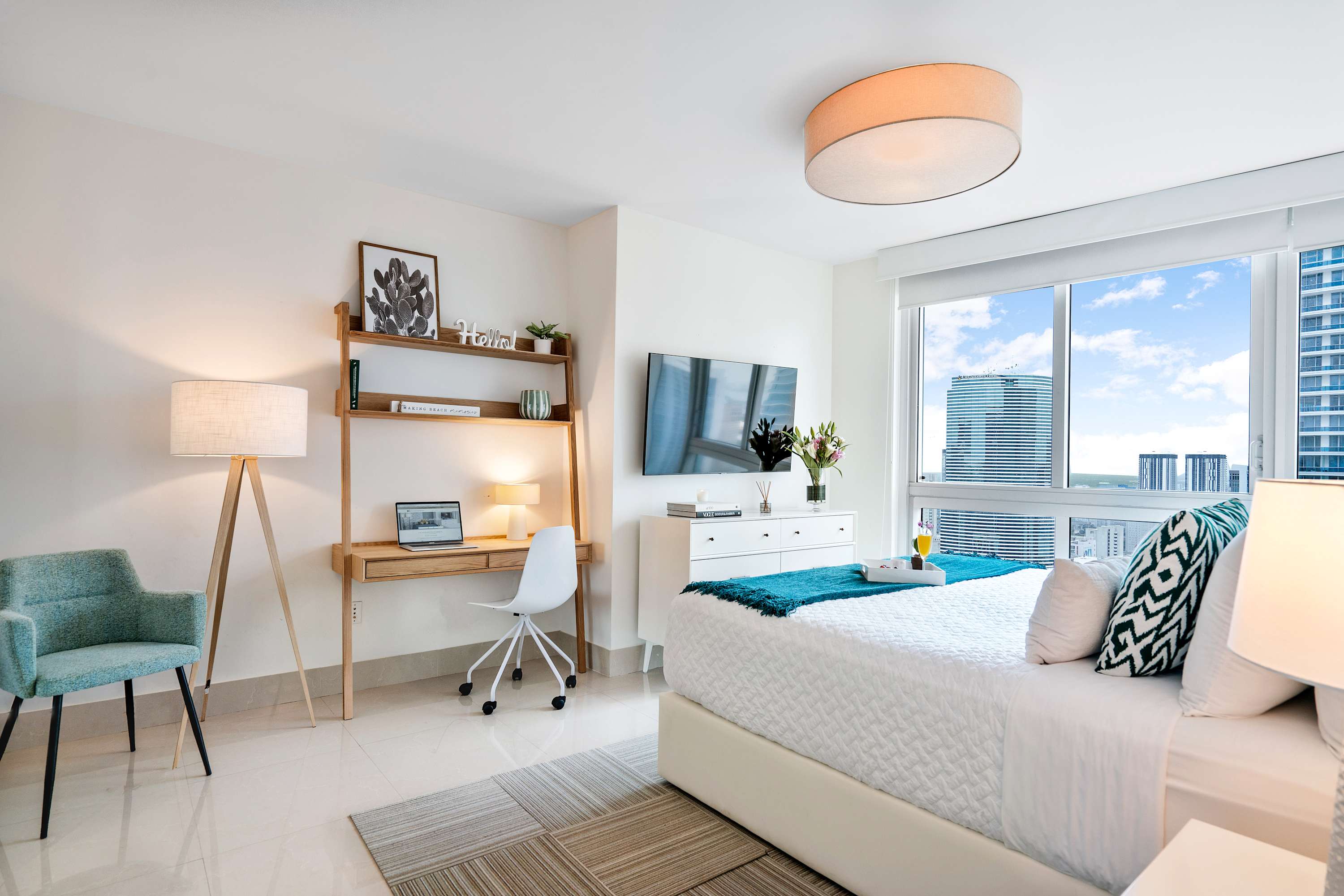 Bright and airy bedroom with a white bed, a dedicated workspace featuring a wooden desk and chair, and large windows offering city views, decorated with a teal accent chair and rug.
