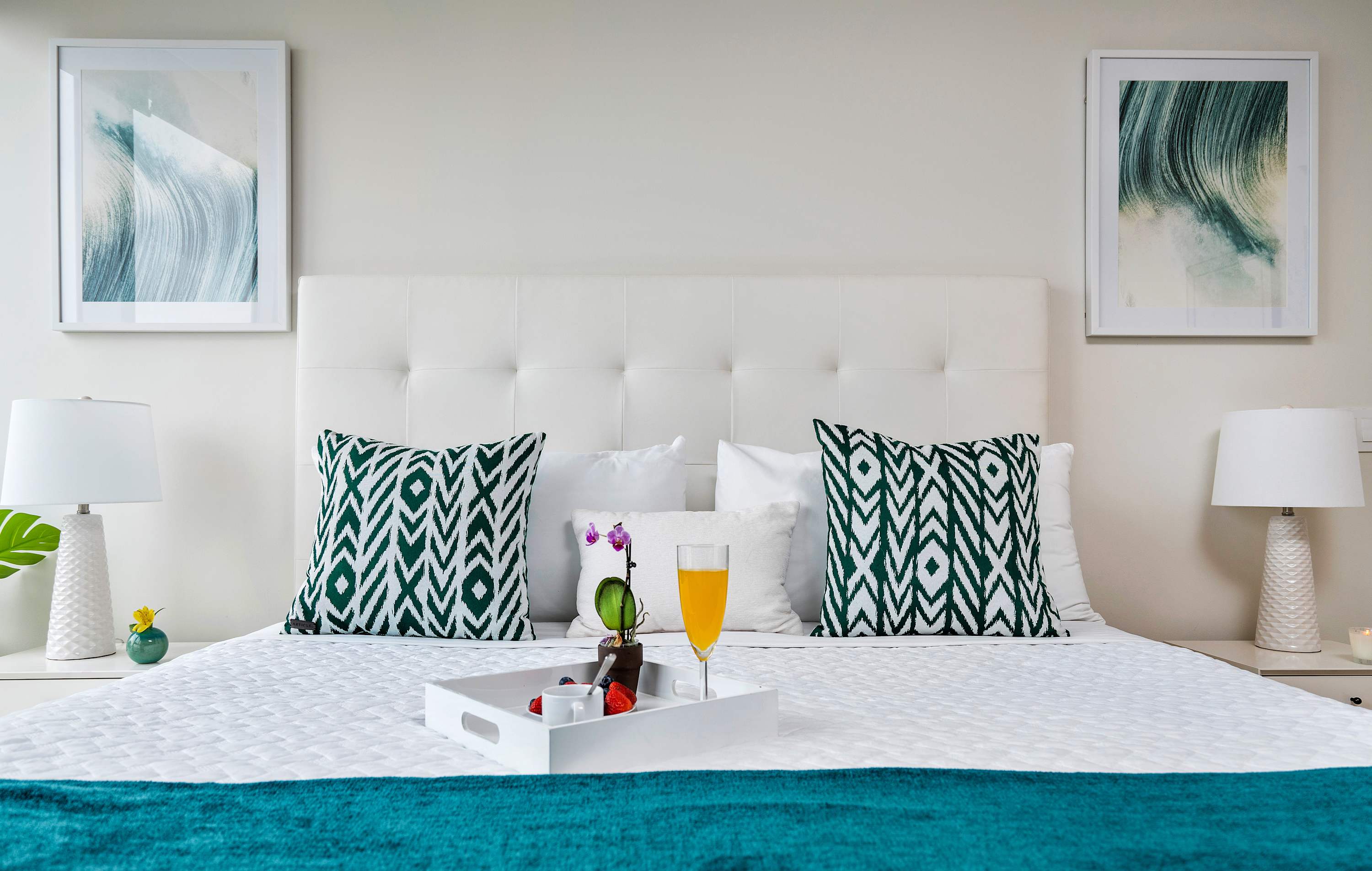 Close-up of a comfortable bed with a white tufted headboard, crisp white bedding, and green patterned pillows, flanked by white nightstands with lamps and abstract artwork above.