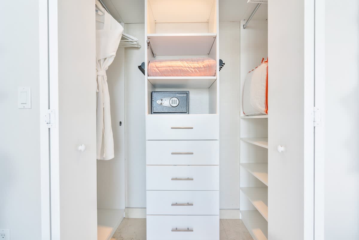 A well-organized white closet features multiple shelves, hanging space, and a central stack of drawers with a small safe. A folded blanket and a laundry bag are visible, indicating storage for guest belongings.