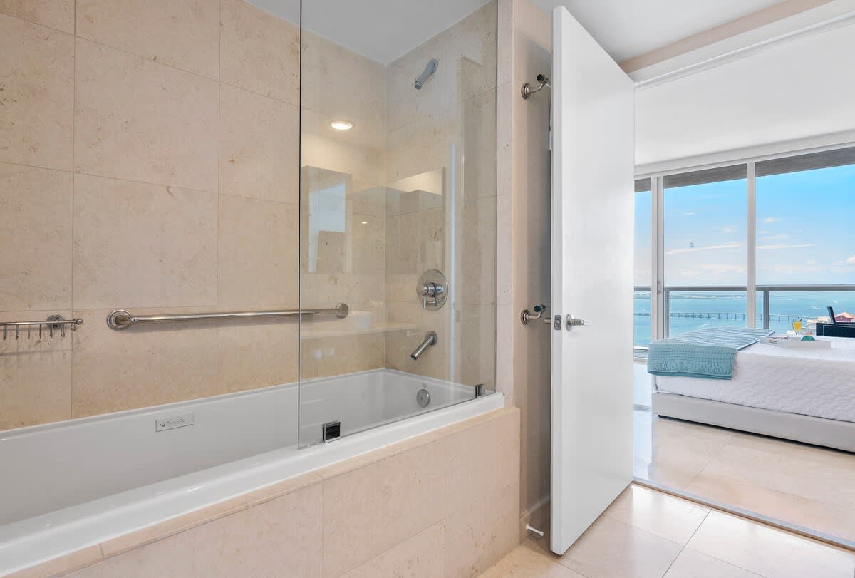 A contemporary bathroom features a built-in bathtub with a glass shower screen and light-colored tiled walls. Chrome fixtures and a grab bar are visible, with a glimpse of a bedroom and a waterfront view through an open doorway.