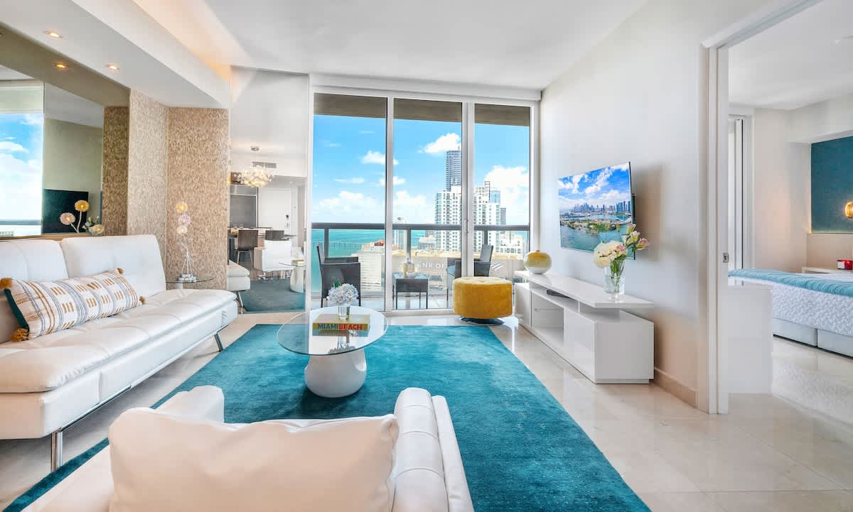 A modern living room with a white sofa, a blue area rug, and a white media console with a flat-screen TV, featuring large windows that provide a clear view of the ocean and city skyline.