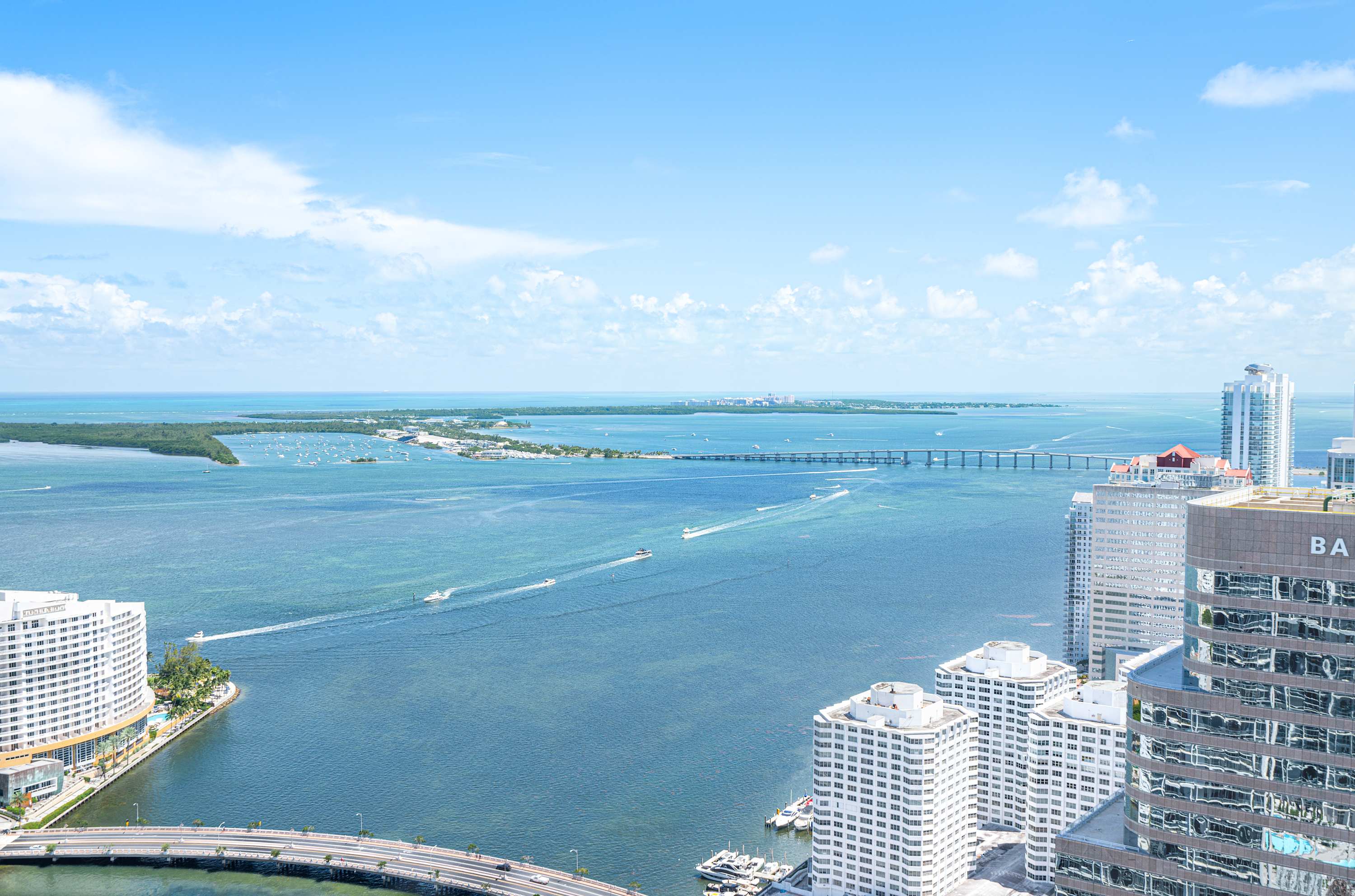 A breathtaking panoramic view of a city skyline and expansive bay from a high vantage point, featuring numerous high-rise buildings, a wide body of water with boats, and distant land under a bright blue sky with scattered clouds.