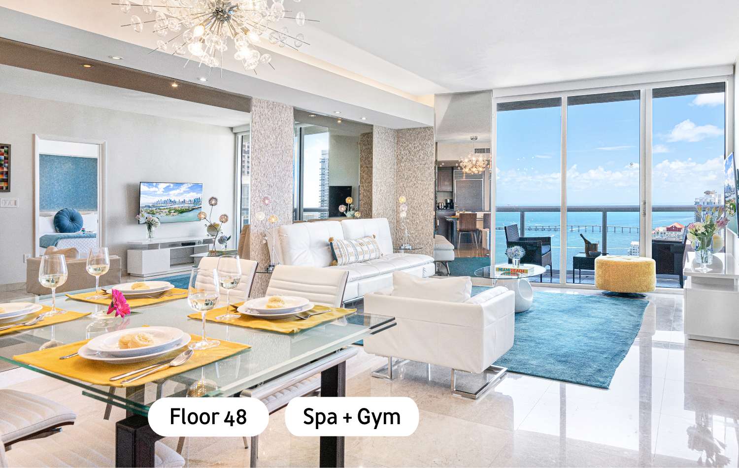 A modern open-concept living and dining area features white leather furniture, a glass dining table with yellow placemats, and large windows offering a panoramic ocean view. The space is illuminated by a sparkling chandelier and has polished light tile floors.