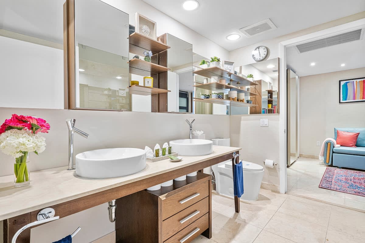 A spacious modern bathroom features a double vanity with two white vessel sinks, chrome faucets, and a light marble countertop over dark wood cabinetry. Large mirrors reflect decorative shelves and a toilet, with a colorful living area visible through the doorway.
