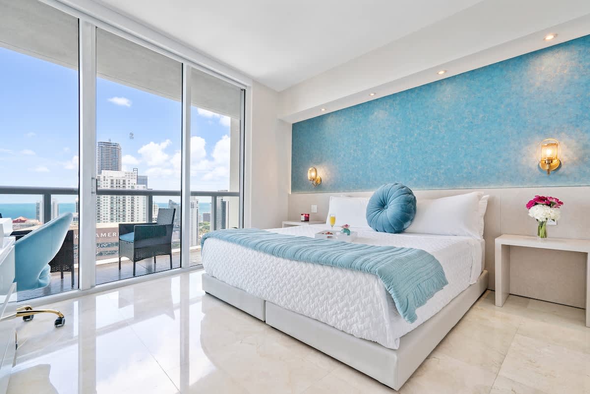 A bright bedroom with a large bed featuring a blue throw and decorative pillow, set against a textured blue accent wall. Floor-to-ceiling windows offer stunning ocean and city views, with a small balcony visible.