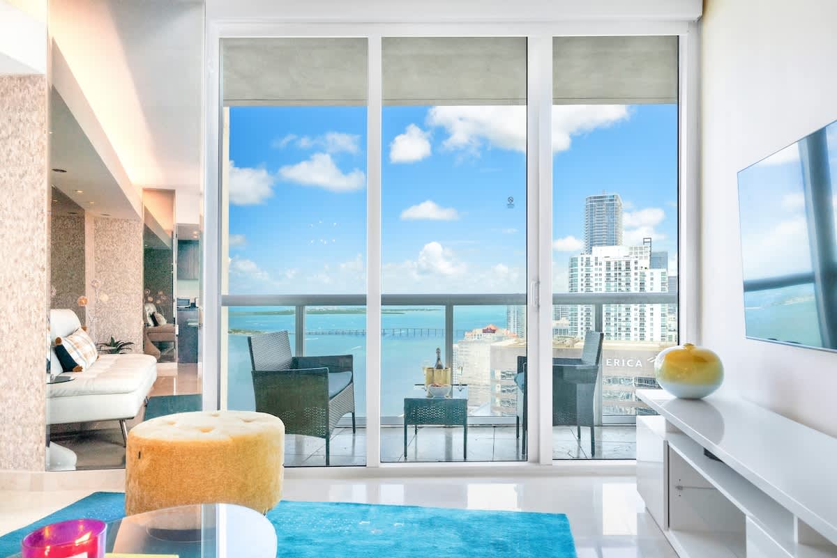 A close-up view of the living room's floor-to-ceiling windows, showcasing the expansive ocean and city views from the high-rise apartment. A glimpse of the white sofa and blue rug is visible inside.