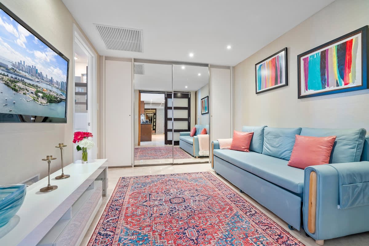 A cozy living room features a light blue sofa with coral throw pillows, a colorful patterned rug, and a wall-mounted TV displaying a city and waterfront view. Mirrored closet doors reflect the room, adding depth and light.