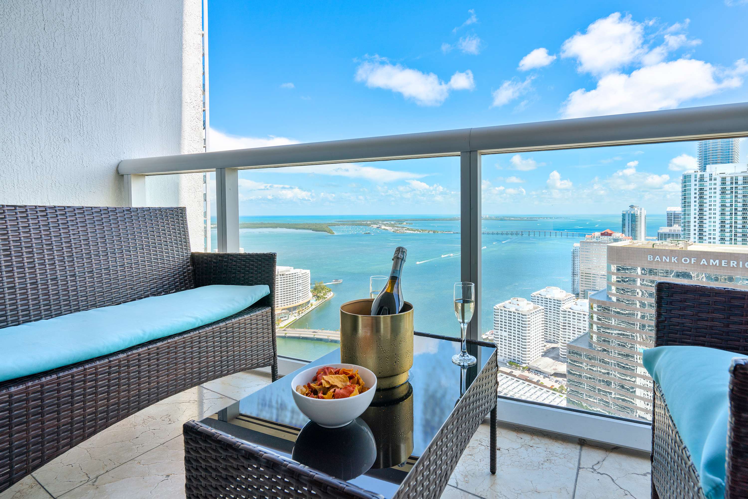 A high-rise balcony with wicker outdoor furniture and a glass table, offering a stunning panoramic view of the ocean, bay, and city skyline under a bright blue sky. A bottle of champagne and snacks are on the table.