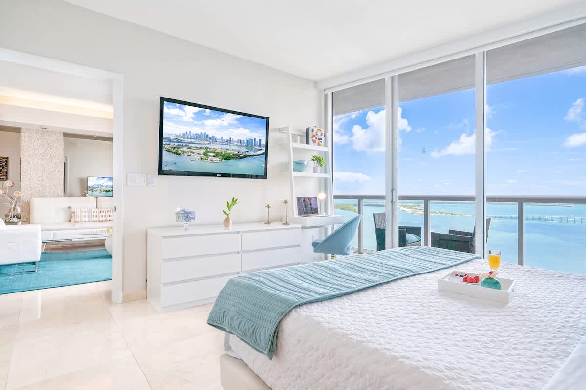 A spacious bedroom with a white bed, a blue throw, and a white dresser with a flat-screen TV mounted above it. Large windows provide panoramic views of the ocean and city, connecting to a living area in the background.
