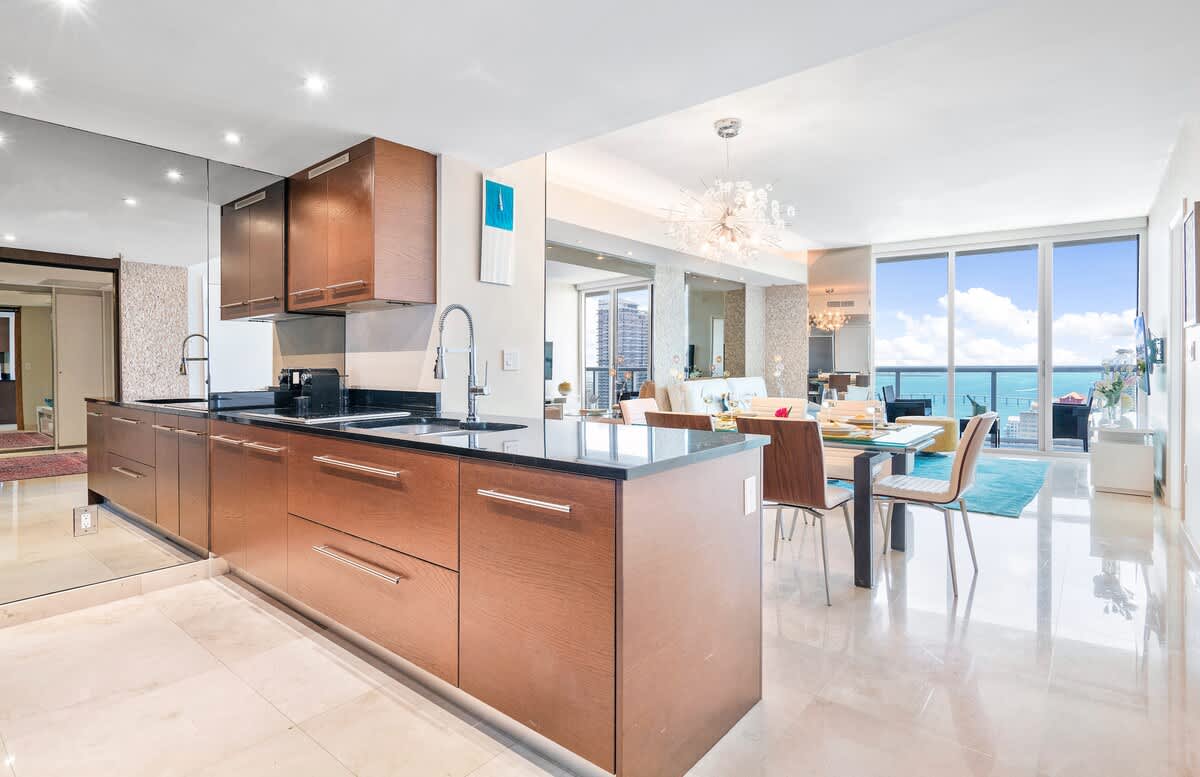 A sleek, modern kitchen with dark wood cabinetry, black countertops, and stainless steel appliances, featuring a large island with a sink and a mirrored backsplash. The kitchen opens to a dining area with a glass table and light wood chairs.