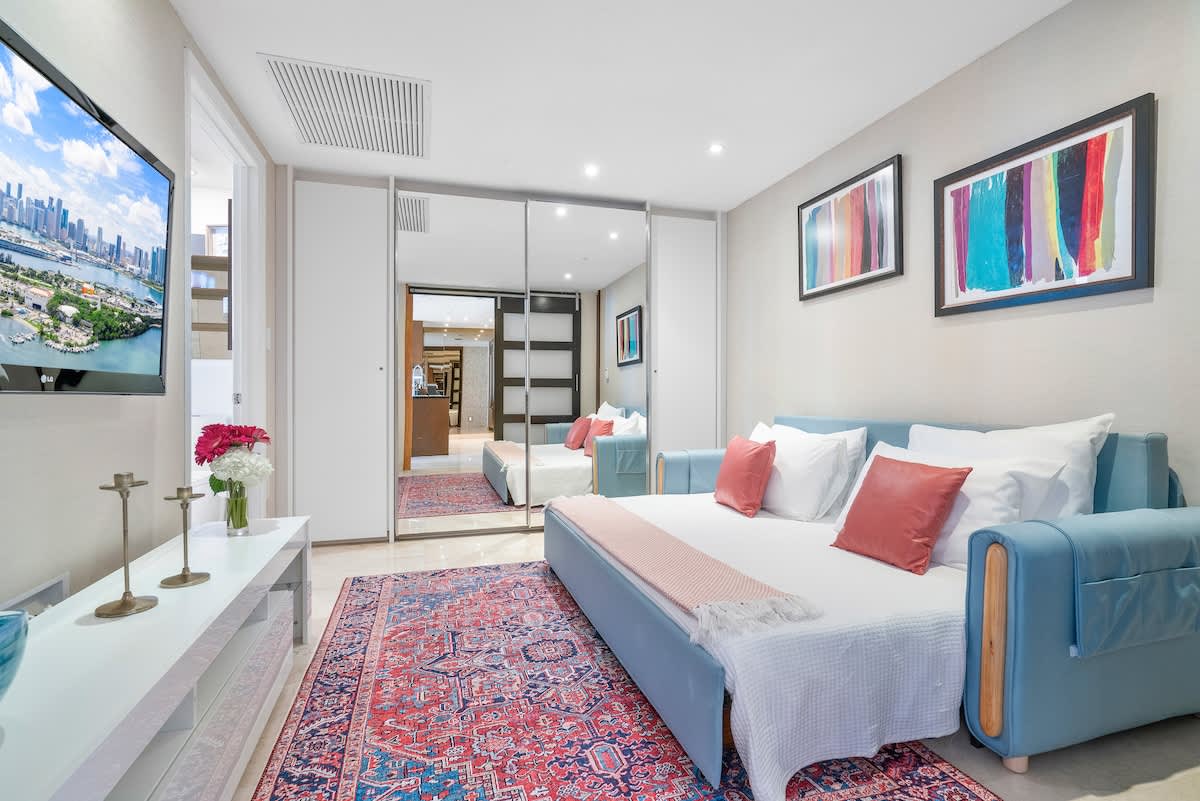 The living room is shown with the light blue sofa converted into a bed, adorned with white bedding and coral throw pillows. A colorful patterned rug covers the floor, and a wall-mounted TV is visible on the left.