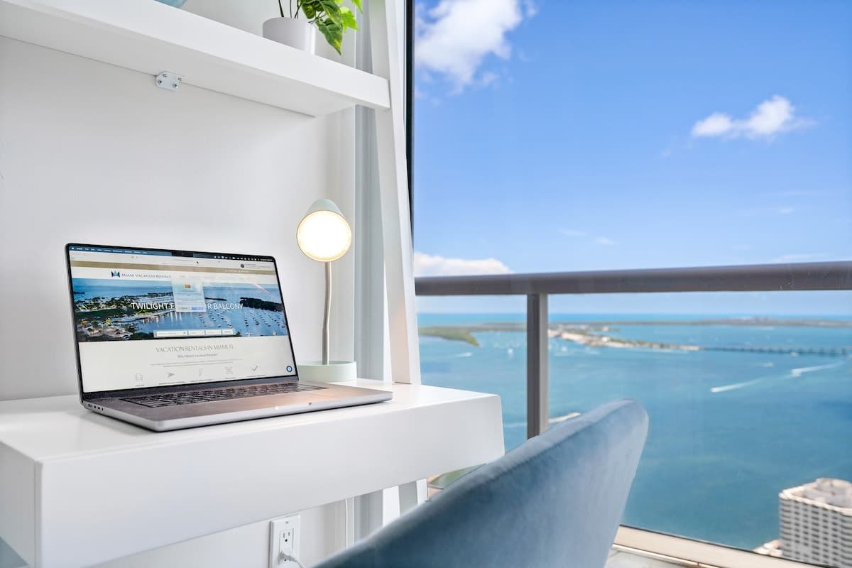 A modern white desk with a laptop and a minimalist lamp is positioned by a large window, offering expansive views of a bay with boats and distant cityscapes under a blue sky. The setup suggests a comfortable workspace with a scenic backdrop.
