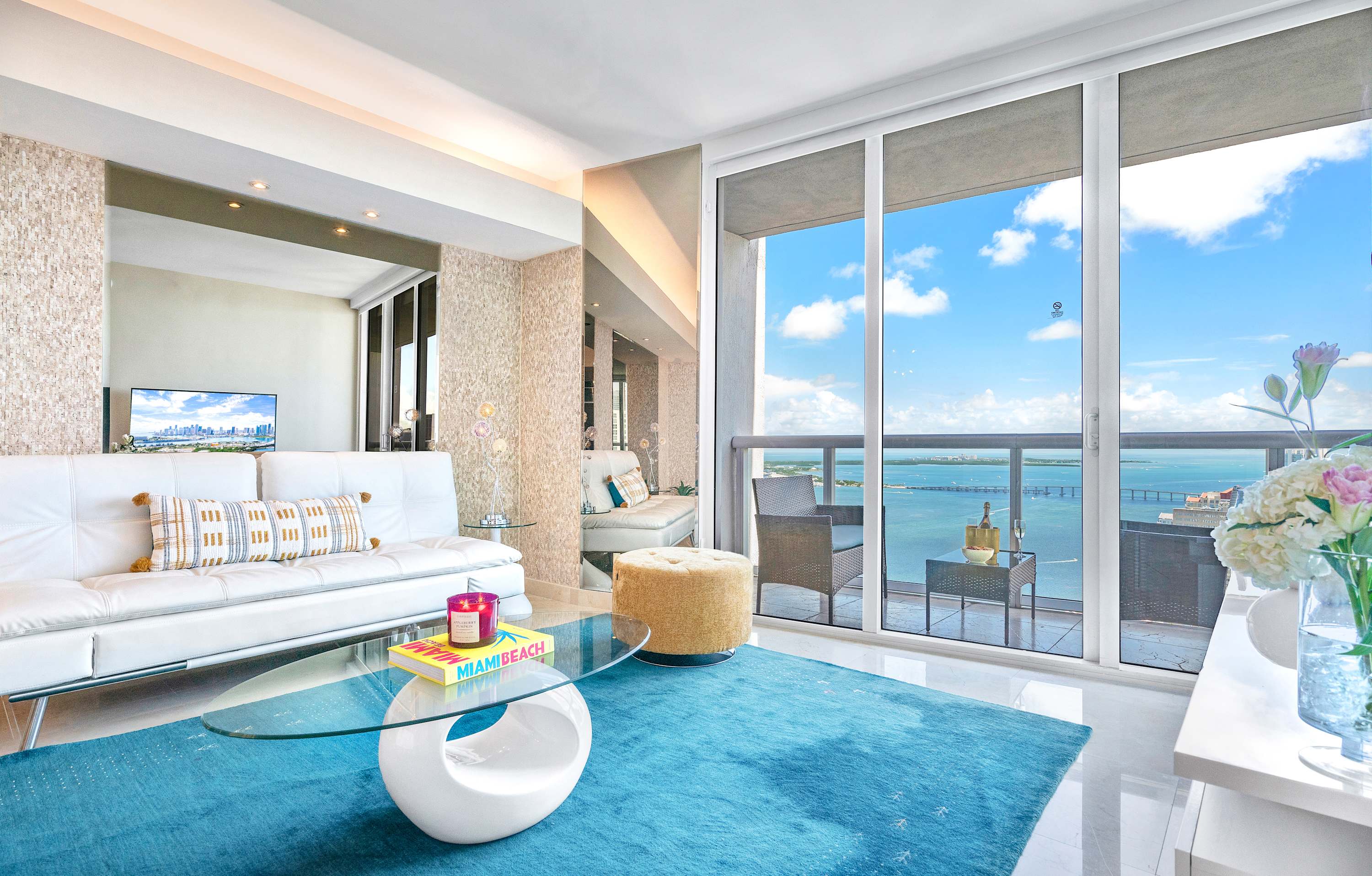 A bright living room with a white leather sofa, a unique white coffee table, and a vibrant blue area rug, featuring floor-to-ceiling windows that open to a balcony with expansive ocean and city views.