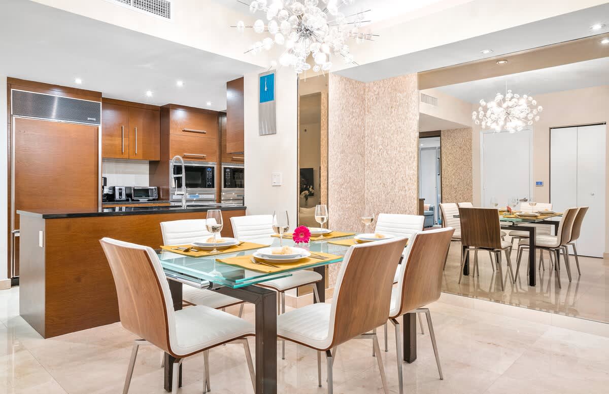 A contemporary dining area with a glass table and six chairs featuring light wood backs and white seats, positioned next to a modern kitchen with dark wood cabinetry. A sparkling chandelier hangs above the table.