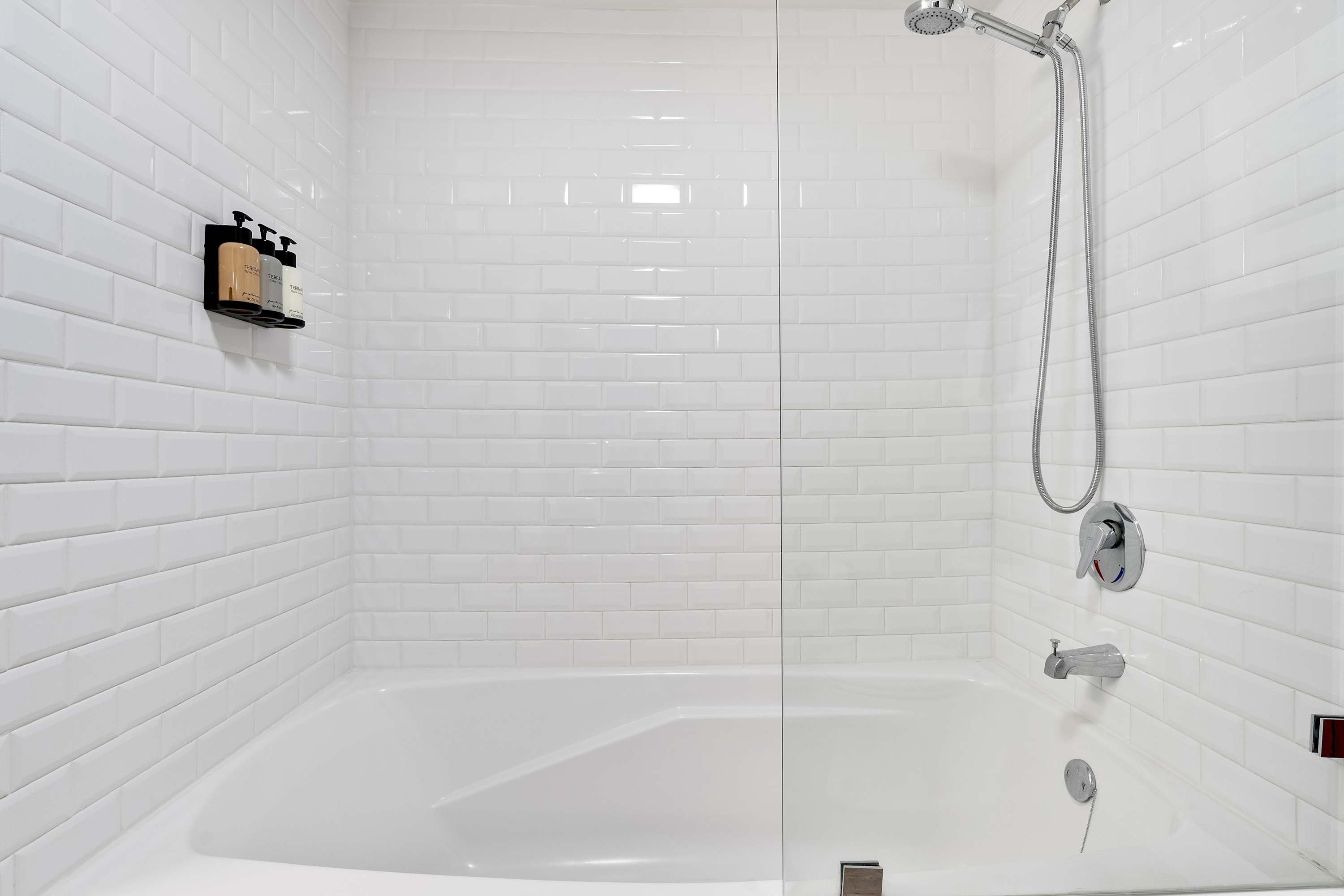 Close-up view of a clean, white bathtub and shower area, featuring white subway tiles, a modern shower head, and bath faucet, with wall-mounted dispensers for toiletries.
