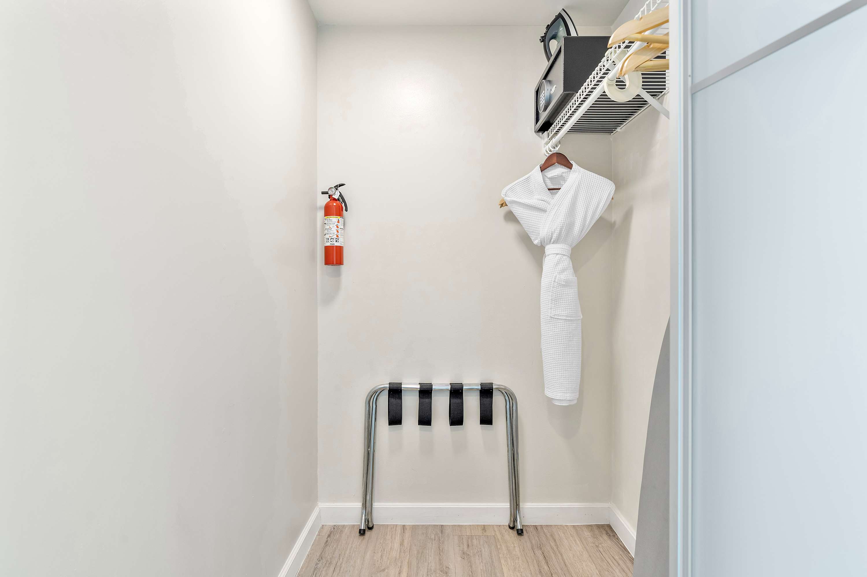 Compact utility closet featuring a fire extinguisher, a metal luggage rack, and a white waffle-knit bathrobe hanging on a shelf with an iron and ironing board.