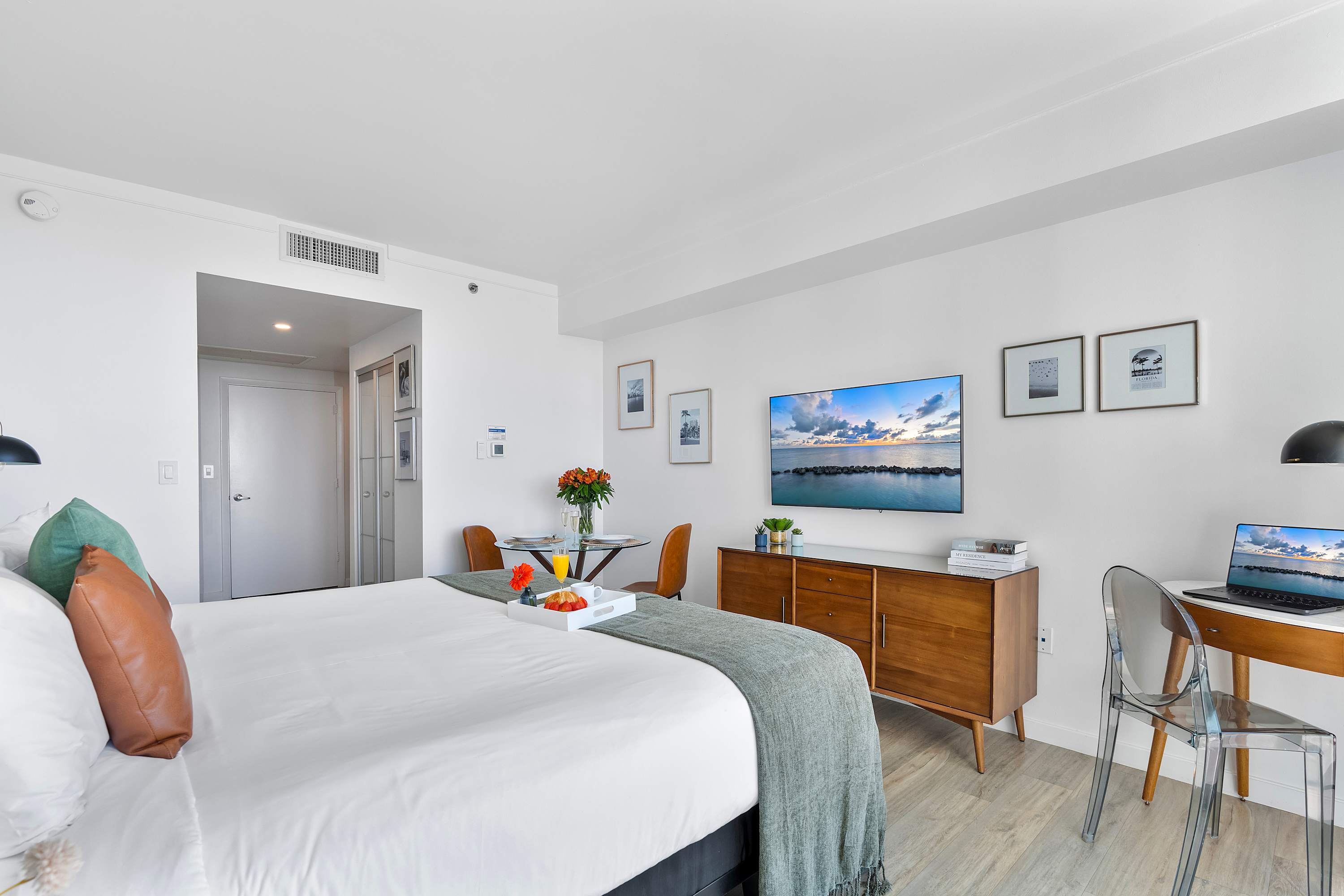 A spacious studio apartment with light wood-look flooring, a large bed, a wall-mounted flat-screen TV, and a glass dining table, all bathed in natural light from the large windows.
