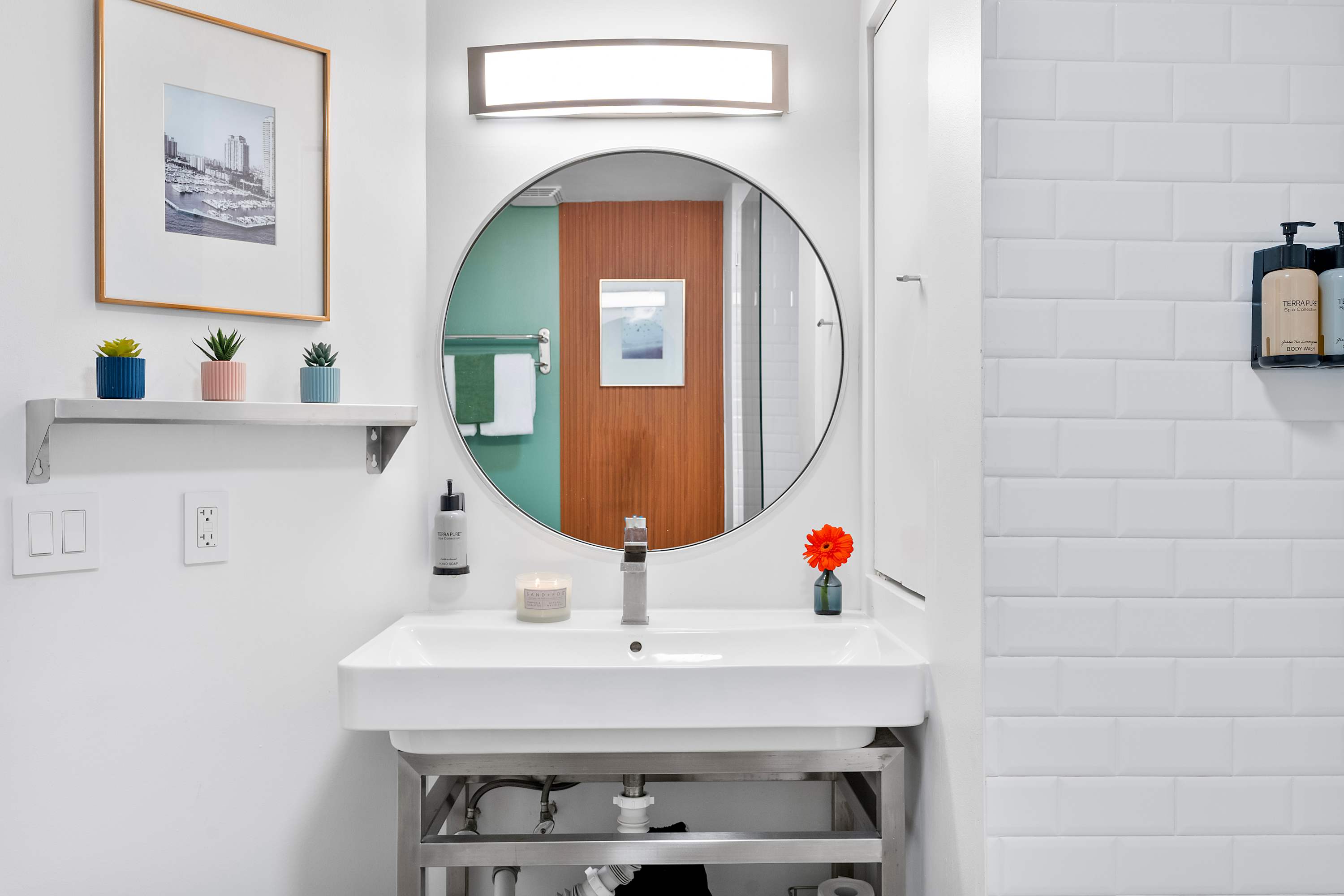 Close-up of a modern bathroom vanity with a white rectangular sink, a large round mirror, and a small shelf adorned with potted succulents, reflecting a warm-toned accent wall.