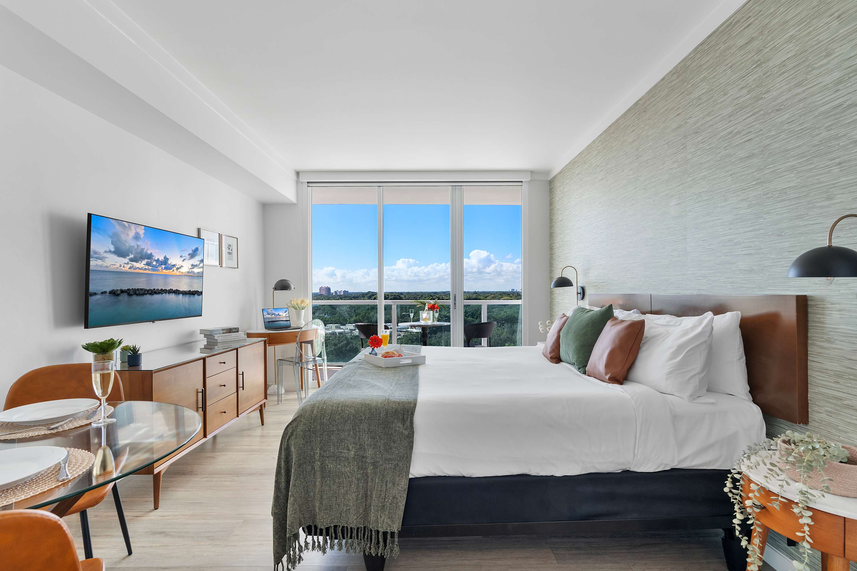 A modern studio apartment showcasing a large bed with a green throw, a small wooden desk with a clear chair, and floor-to-ceiling glass doors leading to a balcony with scenic views.