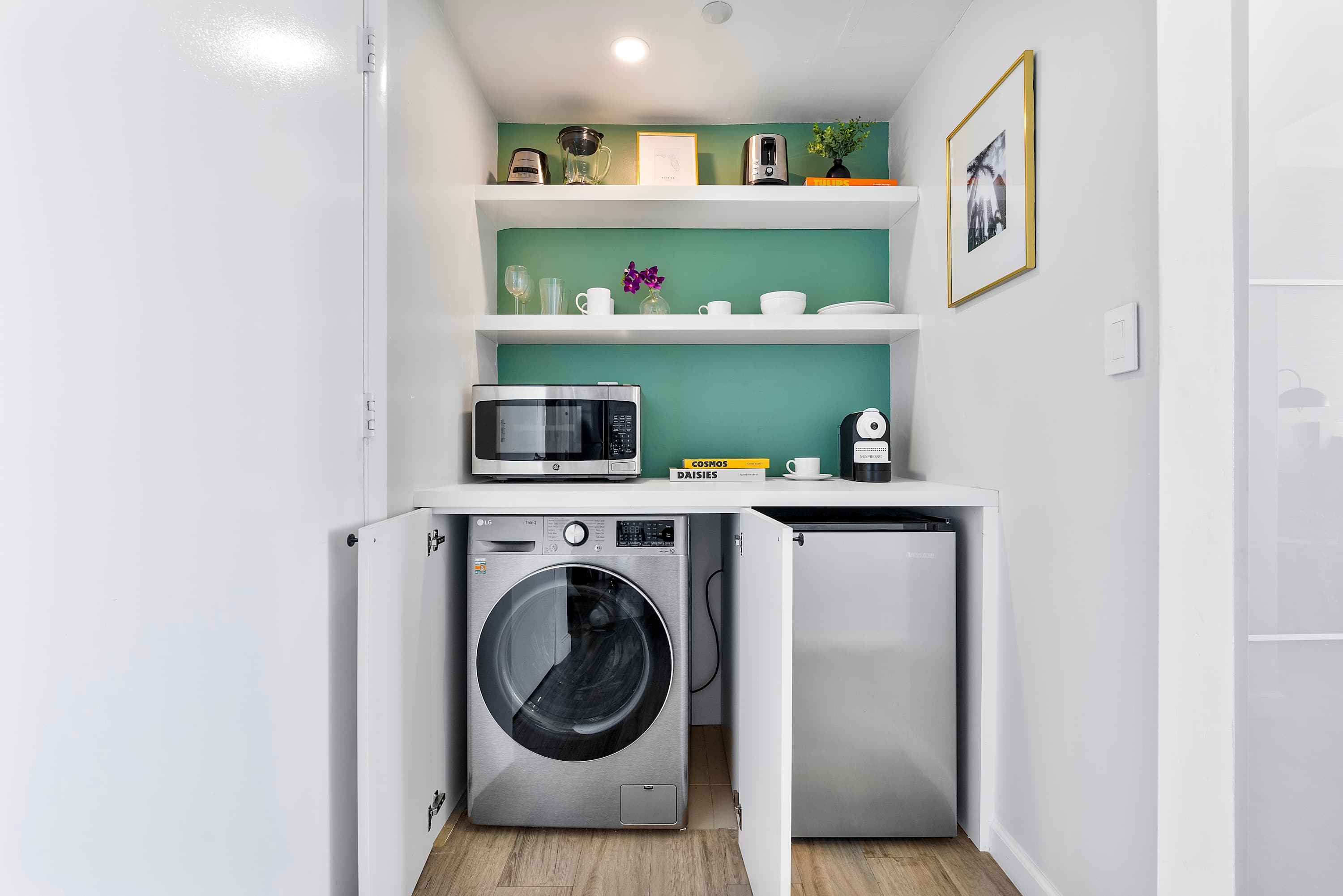 Compact laundry and kitchenette area with a front-loading washing machine and dryer concealed behind cabinet doors, alongside a microwave, mini-fridge, and open shelving with small appliances.