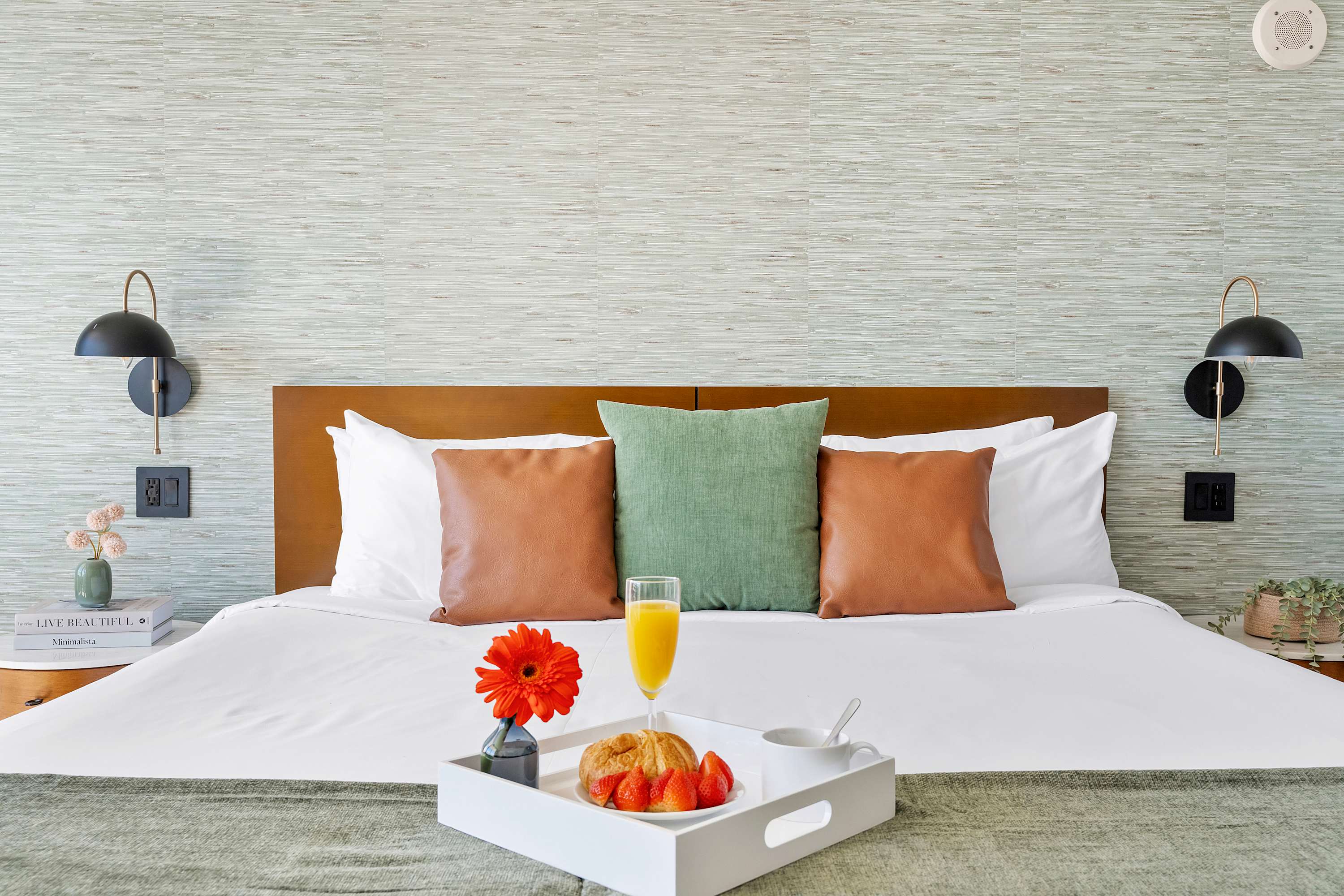 A neatly made bed with white linens, green and brown accent pillows, and a wooden headboard, featuring a breakfast tray with orange juice and a croissant. The wall behind has a textured light green wallpaper.