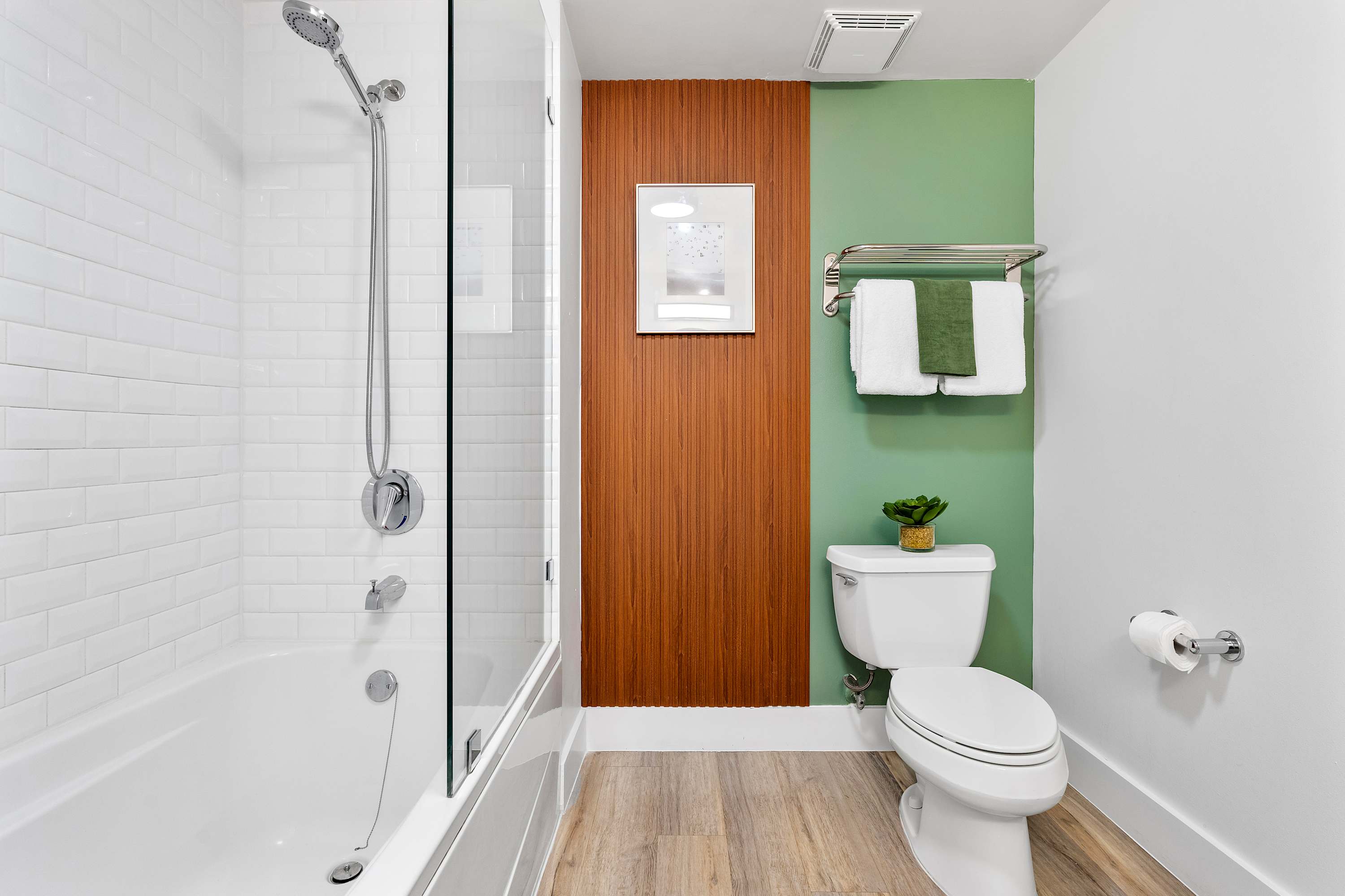 Modern bathroom featuring a white subway-tiled shower/tub combo with a glass partition, a white toilet, and a green accent wall with a wooden panel.