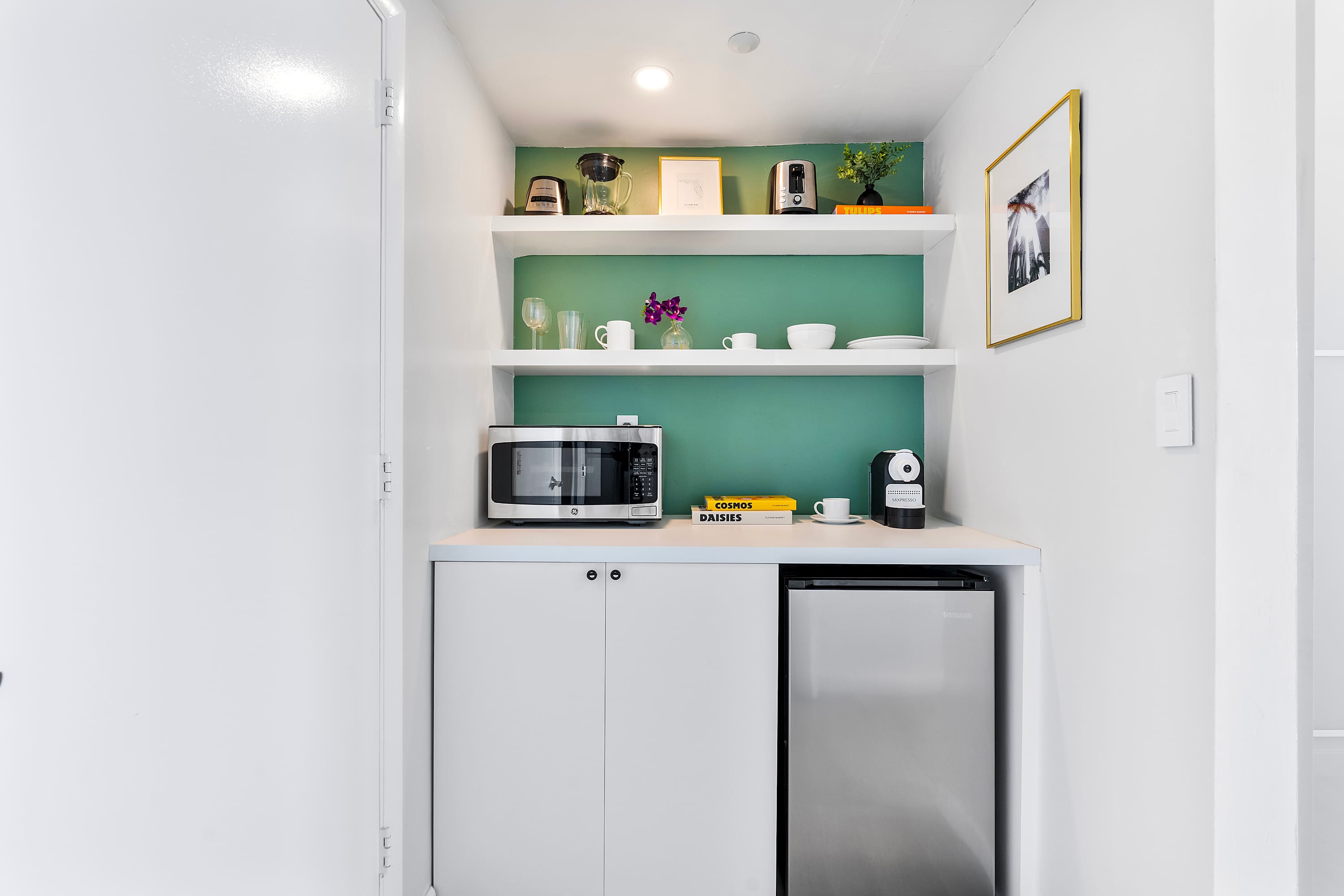 Compact kitchenette area with white open shelving against a light green accent wall, featuring a microwave, mini-fridge, coffee maker, toaster, and various kitchenware.