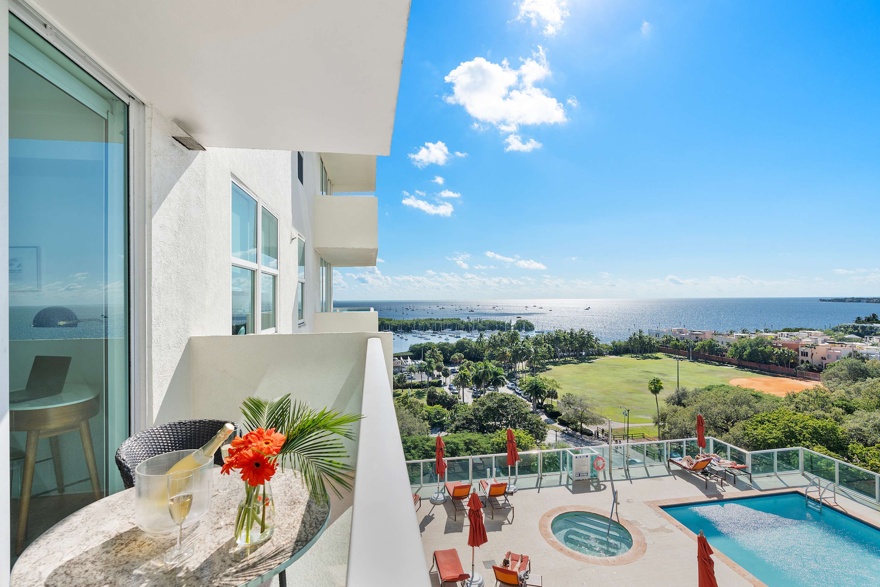 A sunlit balcony with a small outdoor table, offering panoramic views of the ocean, a marina filled with boats, and a resort-style swimming pool with lounge chairs below.
