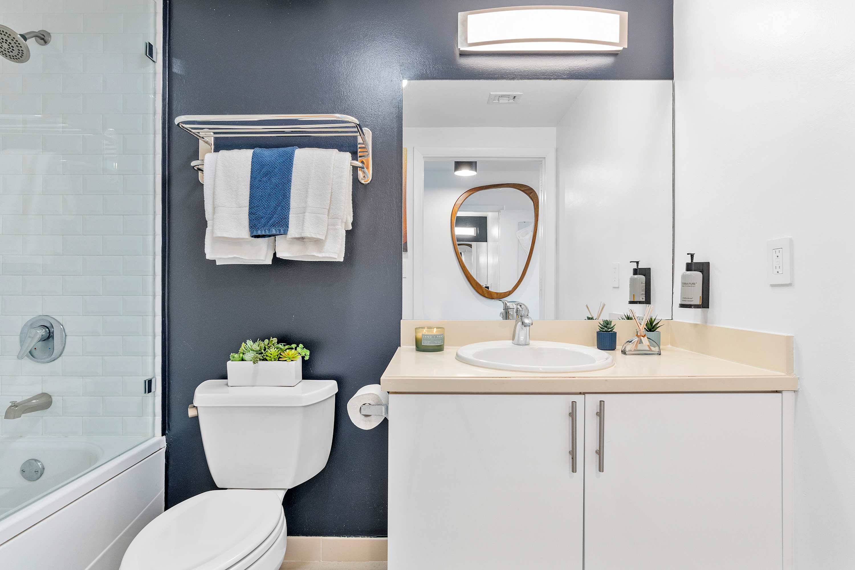 A clean bathroom featuring a white vanity with a sink and mirror, a toilet, and a bathtub with a glass partition. A dark blue accent wall highlights the towel rack.