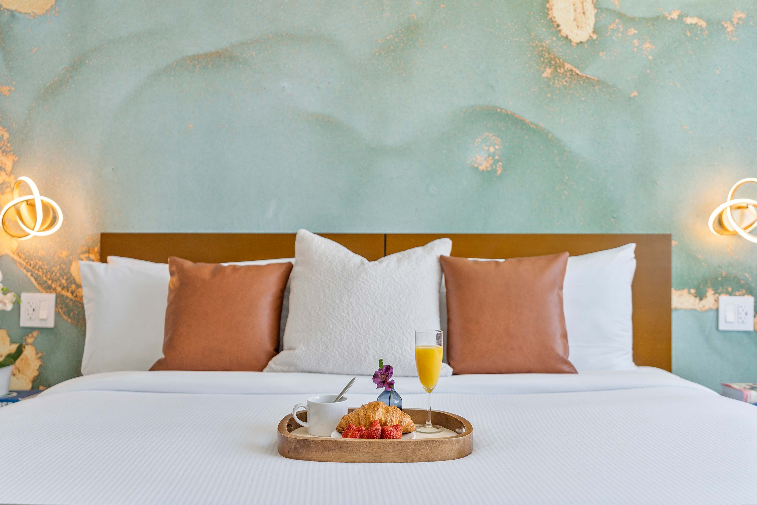 A close-up view of a neatly made bed with white linens, brown leather pillows, and a light blue throw blanket. A wooden tray with breakfast items is placed on the bed.
