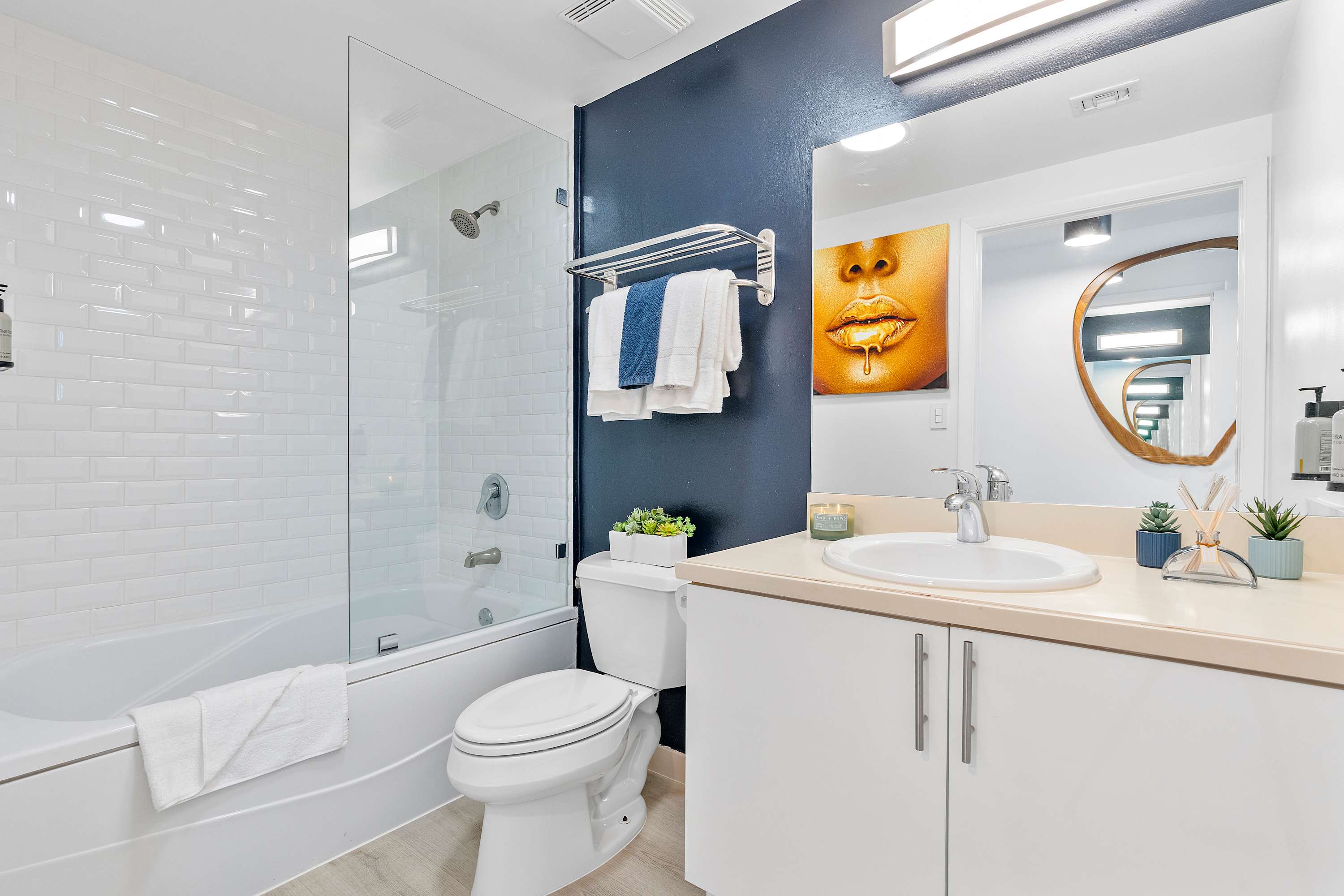 A modern bathroom with a white vanity and sink, a large mirror, a toilet, and a bathtub with a glass shower screen. A dark blue accent wall adds a pop of color.