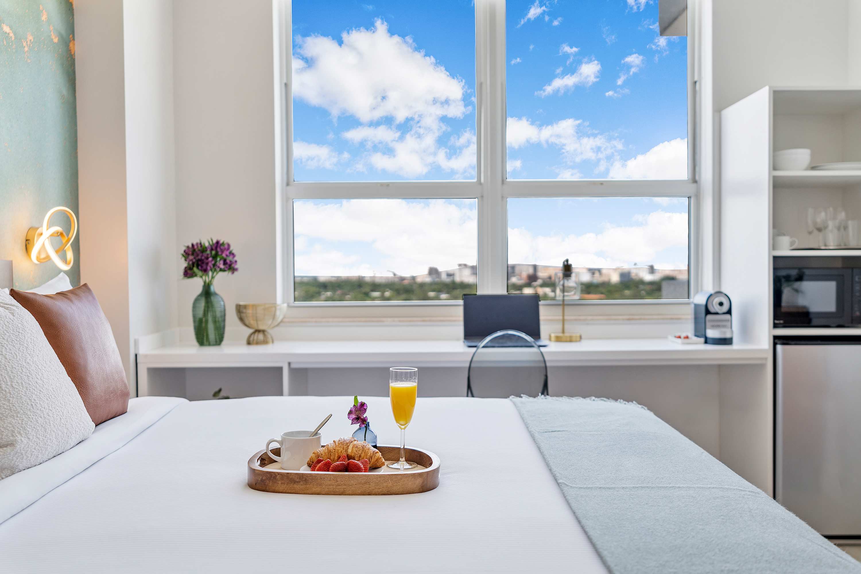 A bright bedroom with a large bed, a long white desk positioned under a wide window with city views, and a breakfast tray on the bed. A laptop and coffee machine are on the desk.