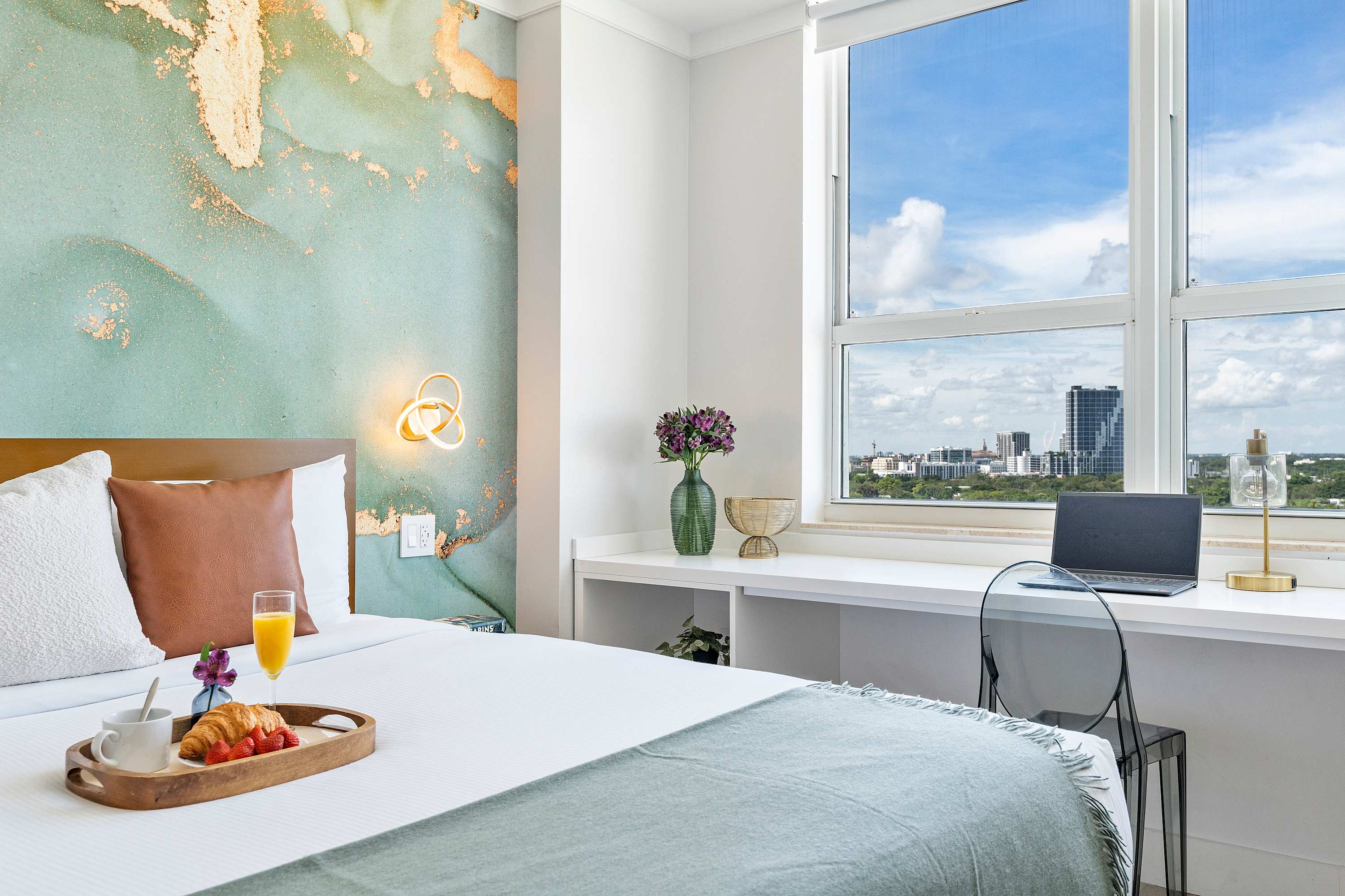 A modern bedroom with a large bed, a distinctive light blue and gold accent wall, and a white desk with a clear chair positioned by a large window offering city views.
