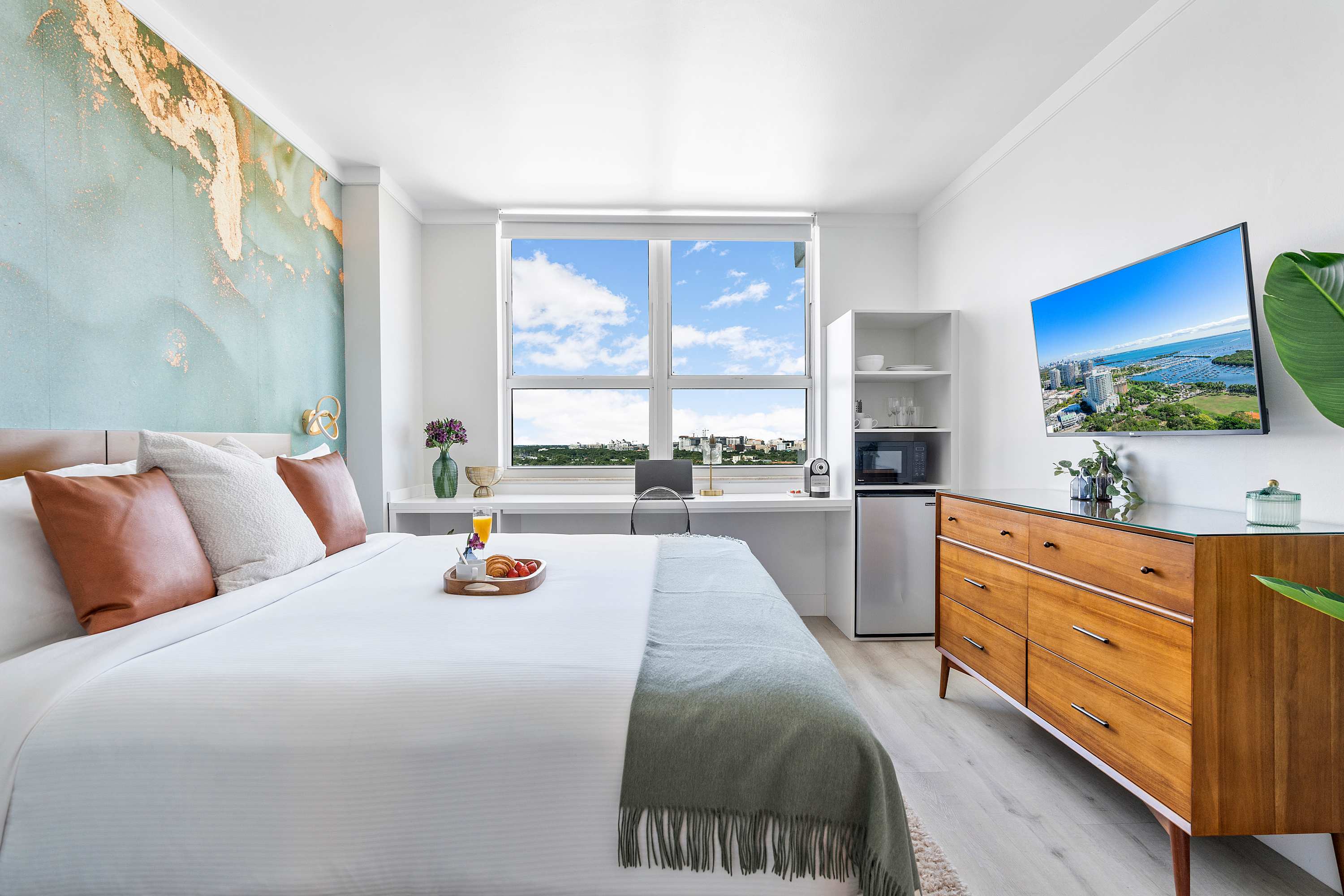 A spacious bedroom featuring a large bed with a breakfast tray, a long white desk beneath a wide window showcasing city views, and a wall-mounted TV above a wooden dresser.