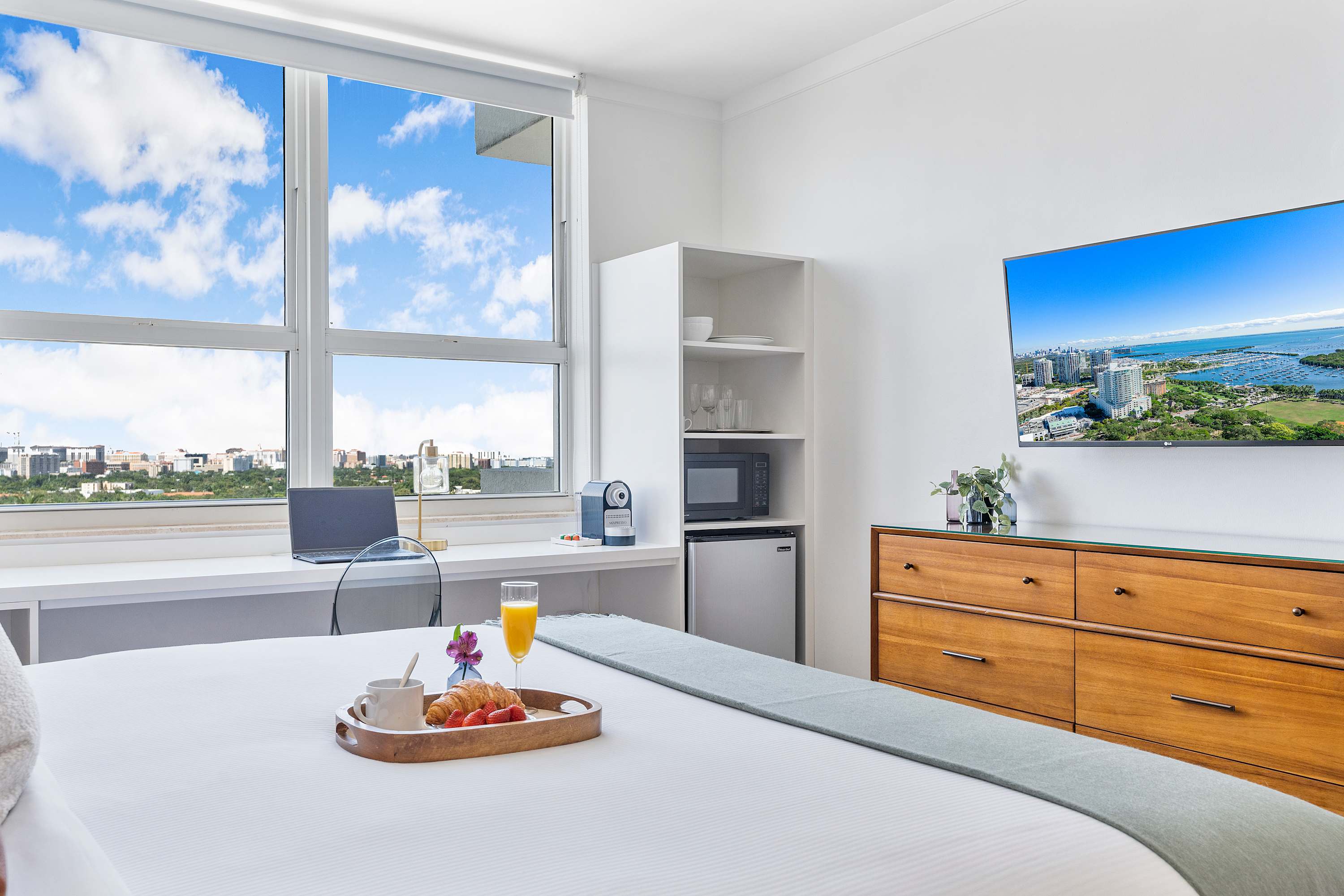 A bright bedroom featuring a large bed with a breakfast tray, a long white desk beneath a wide window showcasing city views, and a wall-mounted TV opposite the bed. A mini-fridge and microwave are visible.