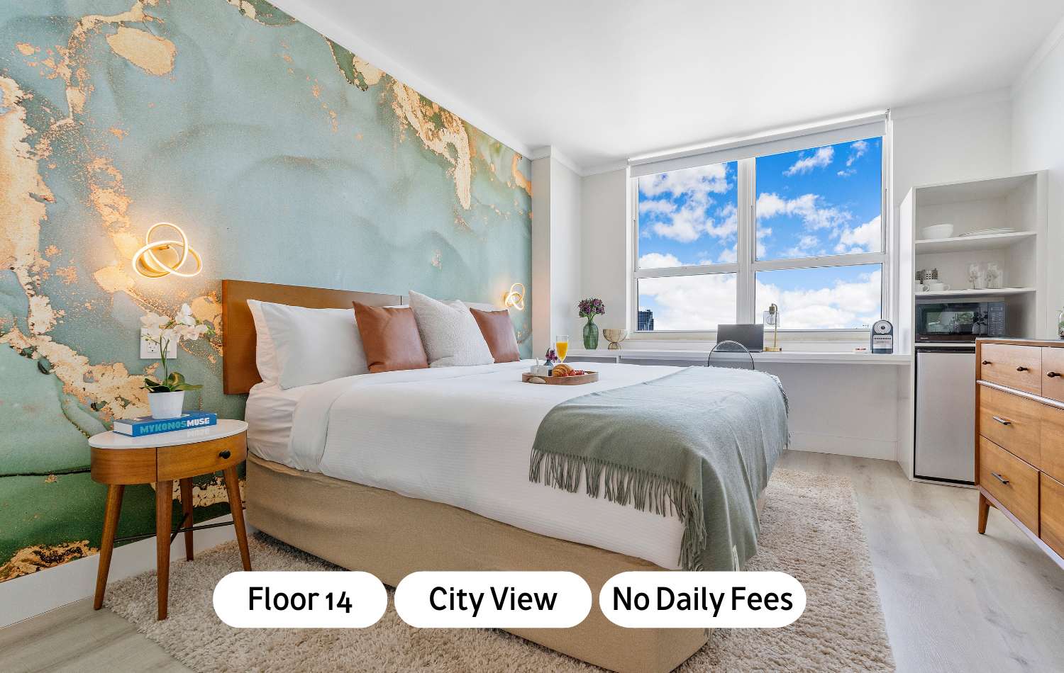 A modern bedroom with a large bed, a light blue and gold abstract accent wall, and a desk by a large window offering city views. A breakfast tray is on the bed.