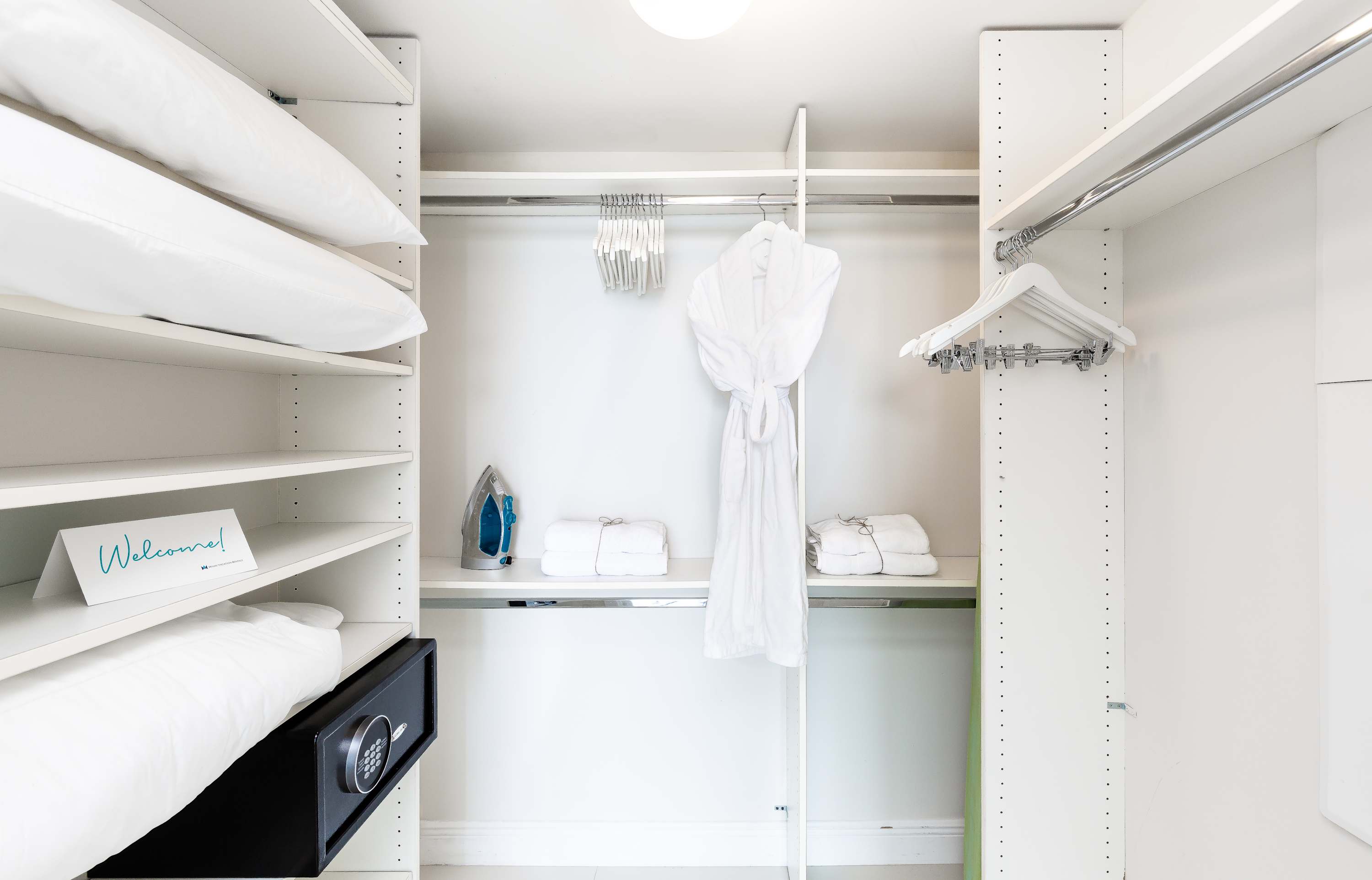 A well-organized white closet with shelving, hanging space, and a small safe. It contains neatly folded towels, a white bathrobe, an iron, and extra pillows.